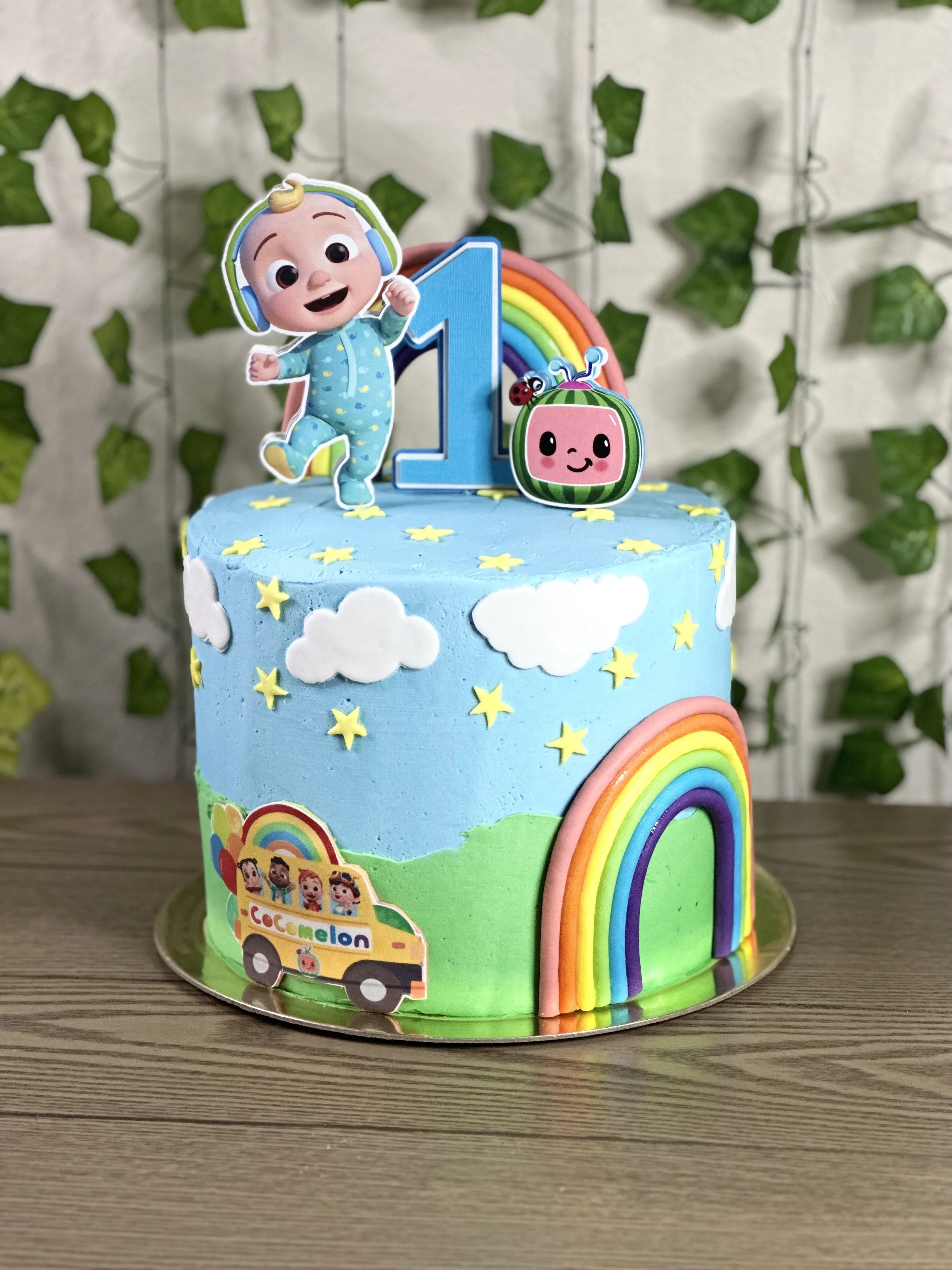 Colorful first birthday cake with a blue sky, clouds, stars, green hills, rainbows, and characters from CoComelon, topped with a cartoon baby figure, a number one, and a rainbow with a ladybug.