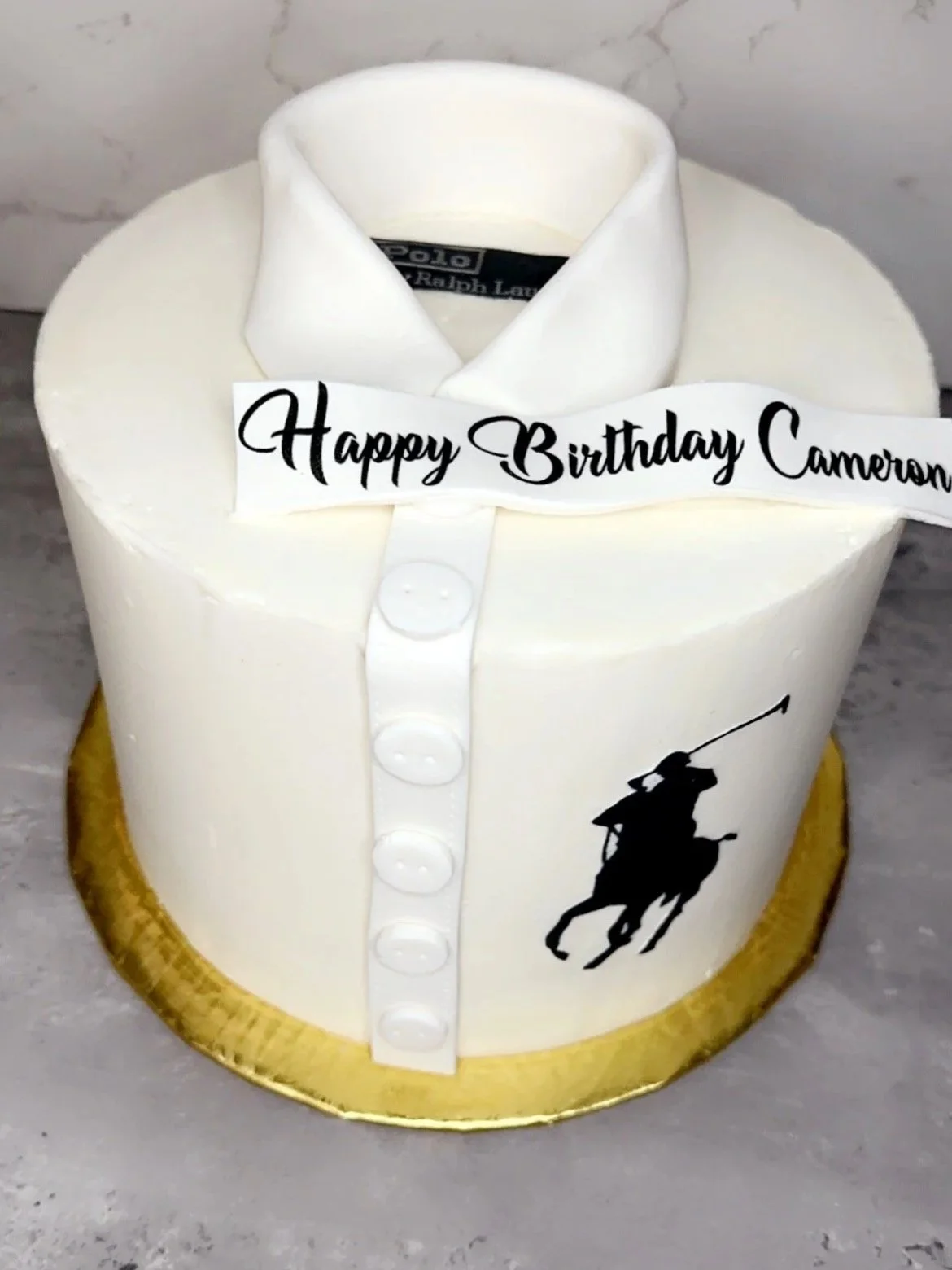 Cake designed to look like a white dress shirt with a Black Polo Ralph Lauren logo on the side. It has a string of buttons down the front, a collar, and a ribbon on top that reads 'Happy Birthday Cameron'.