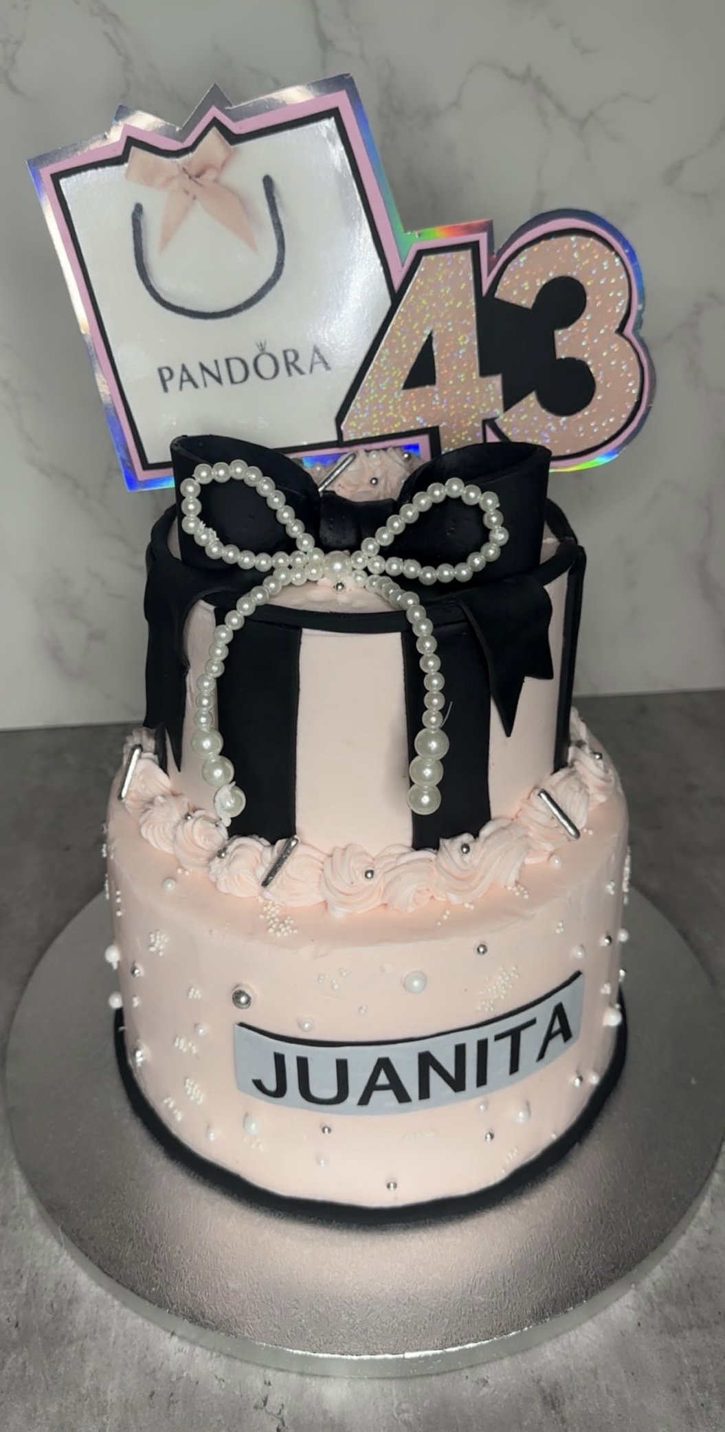 A two-tiered birthday cake decorated with pink frosting, pearl-like beads, and black fondant ribbons, with a pearl bow on top. The cake has a sign with the name "Juanita" and two toppers: a Pandora shopping bag and the number 43 glittery decoration, set against a marble background.