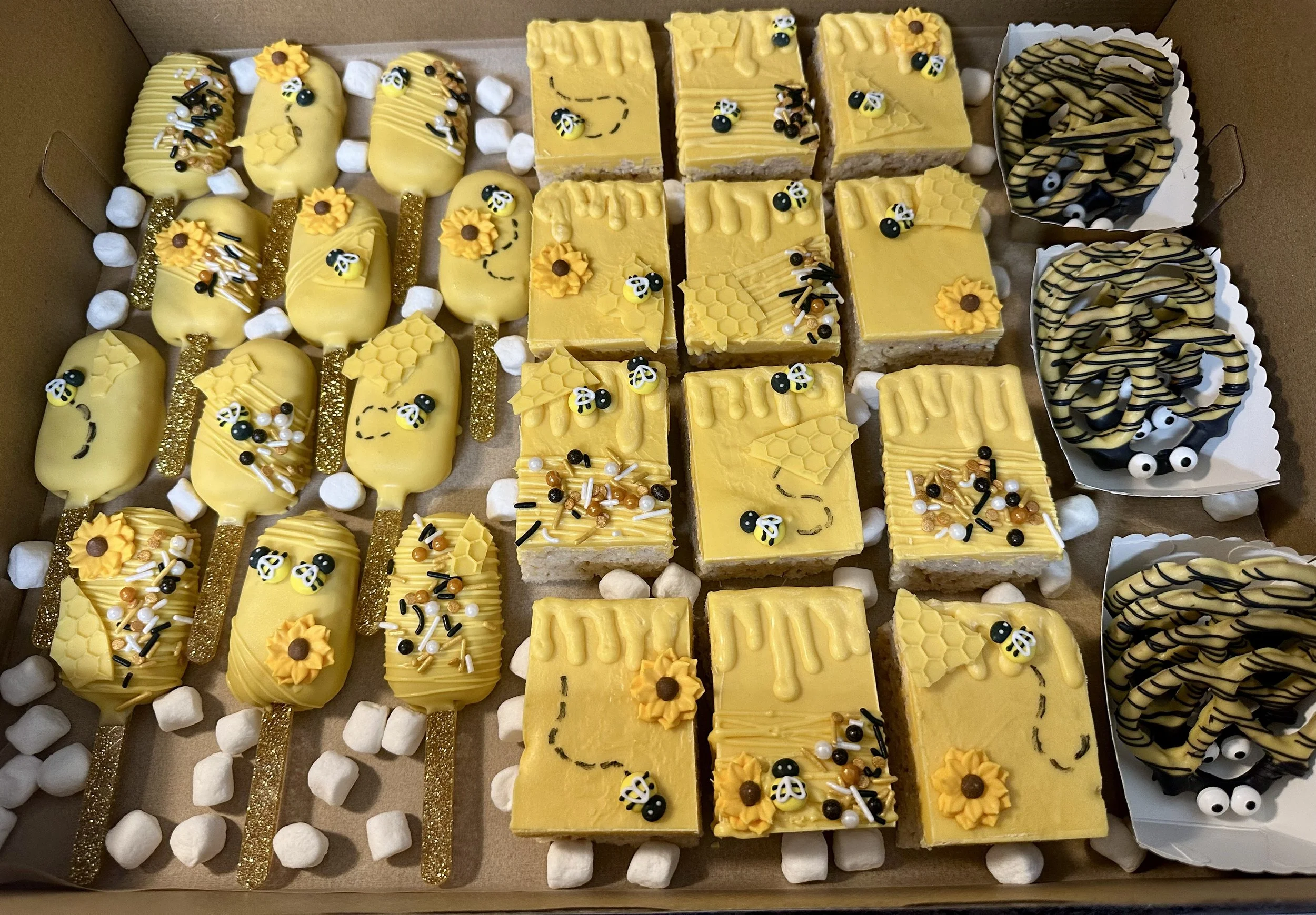 A box filled with yellow bee-themed desserts including cake pops, small square cakes, and chocolate-covered pretzels decorated with bee and honeycomb designs, topped with edible bee and flower decorations.
