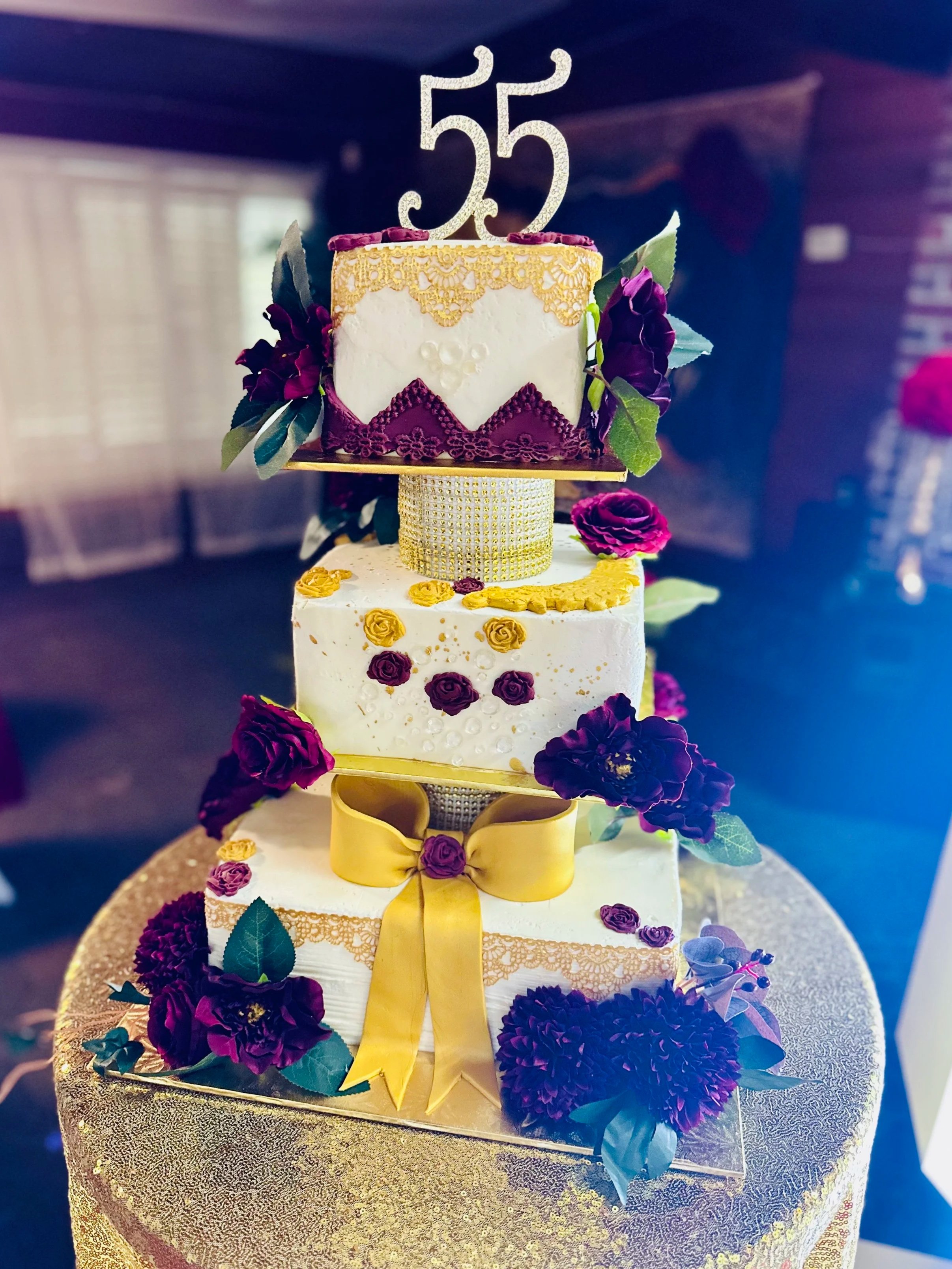 A multi-tiered cake decorated with purple and yellow flowers, a gold bow, and a gold "55" topper, placed on a gold tablecloth.