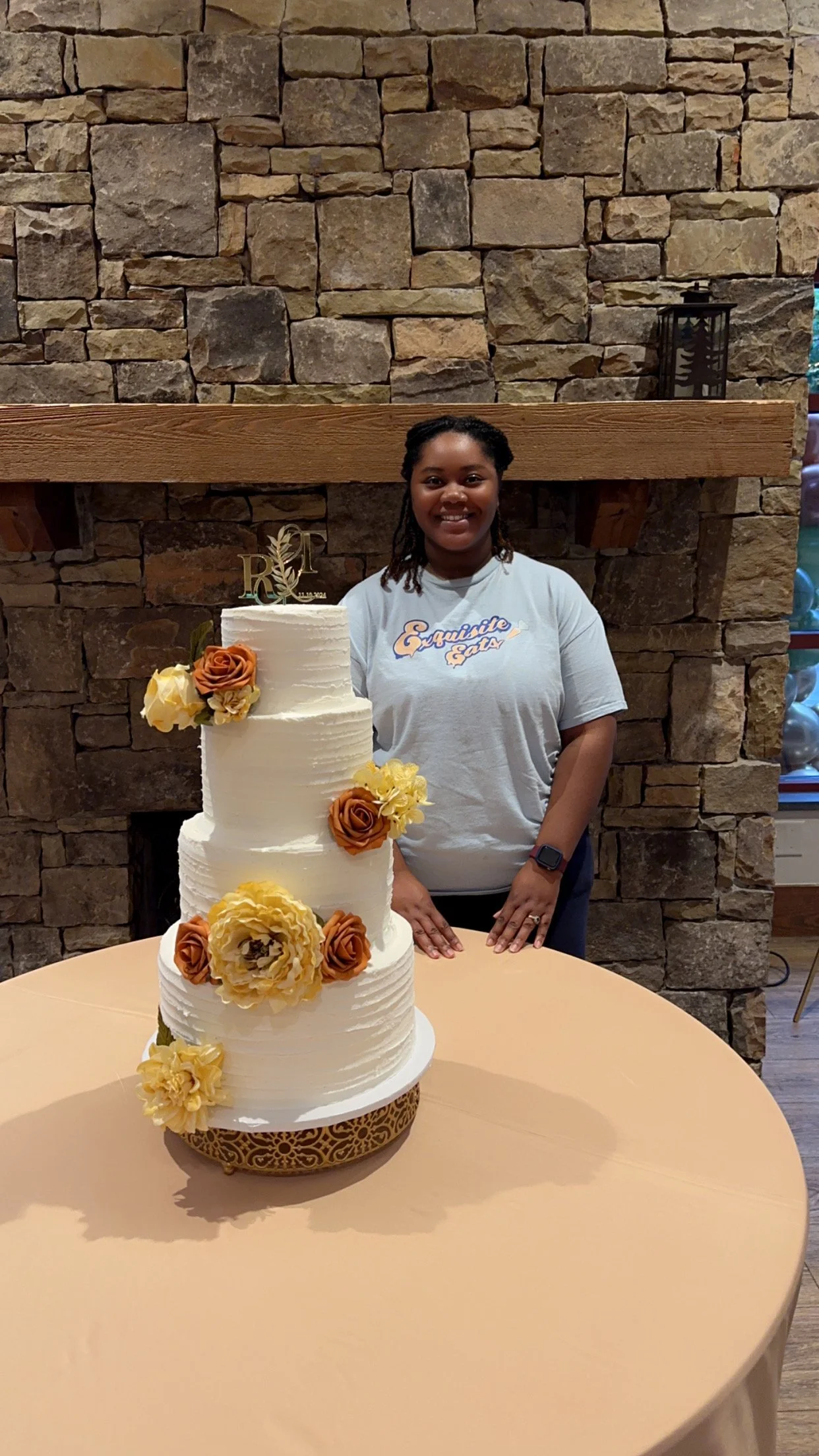 A woman standing behind a three-tier white wedding cake decorated with orange and yellow flowers and a gold cake topper, in front of a stone fireplace.