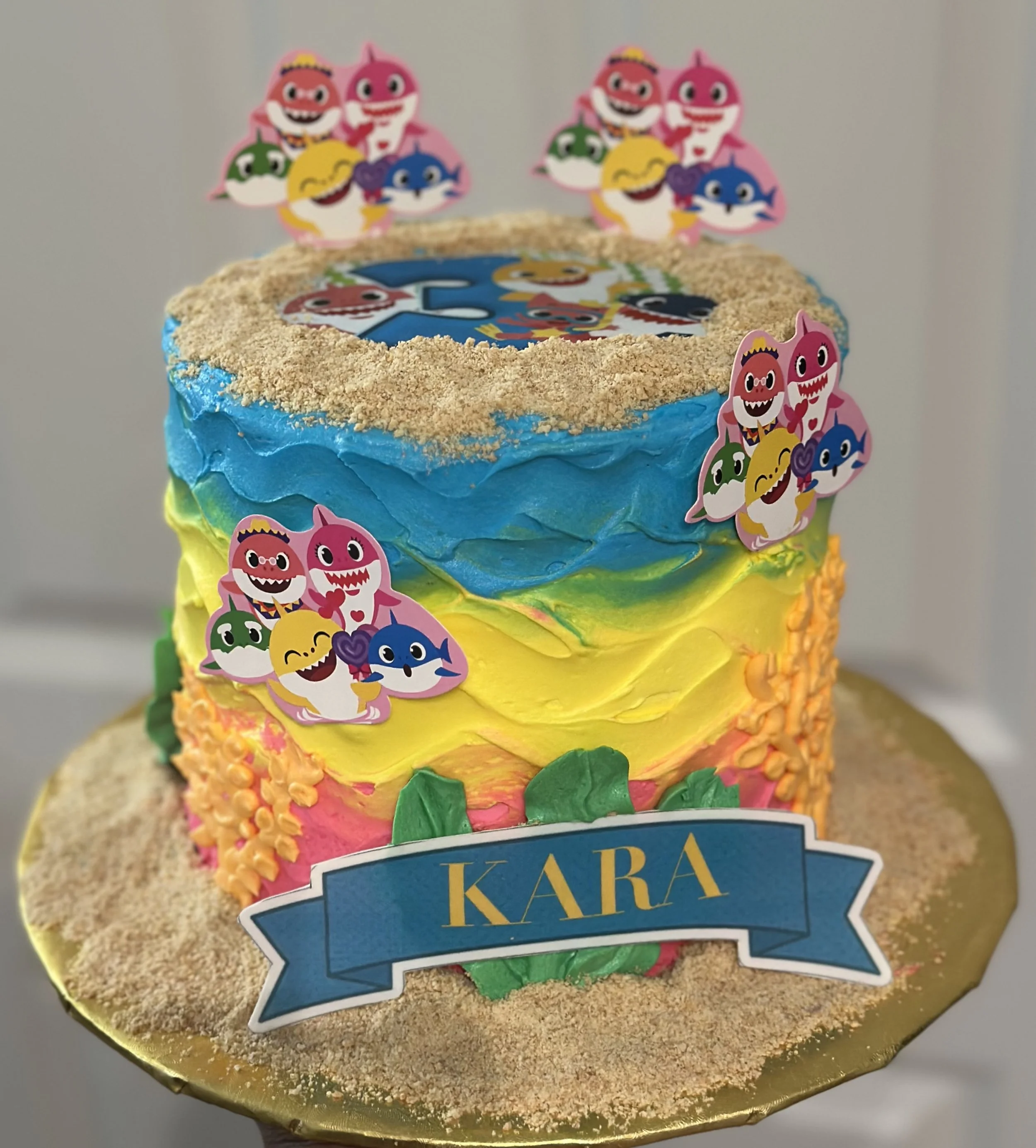 Colorful birthday cake with rainbow-colored frosting, topped with edible cartoon character decorations from the Trolls series, and a blue banner with the name 'KARA' written in yellow.