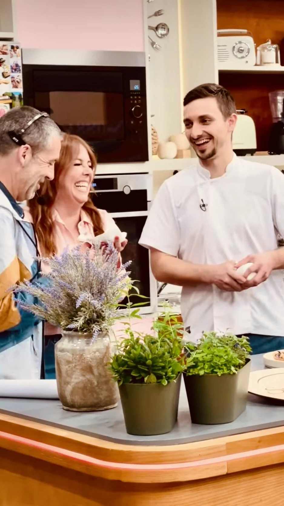Three people laughing and talking in a kitchen, one person is a chef holding eggs, two others are a man and woman. There are potted herbs and a vase of lavender on the kitchen counter.