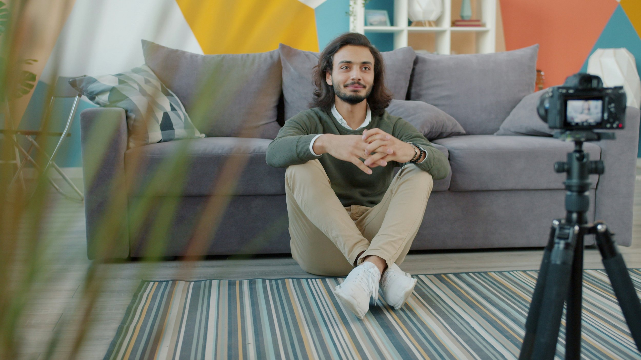 A man sitting on the floor in front of a camera set up on a tripod, inside a colorful and modern living room.