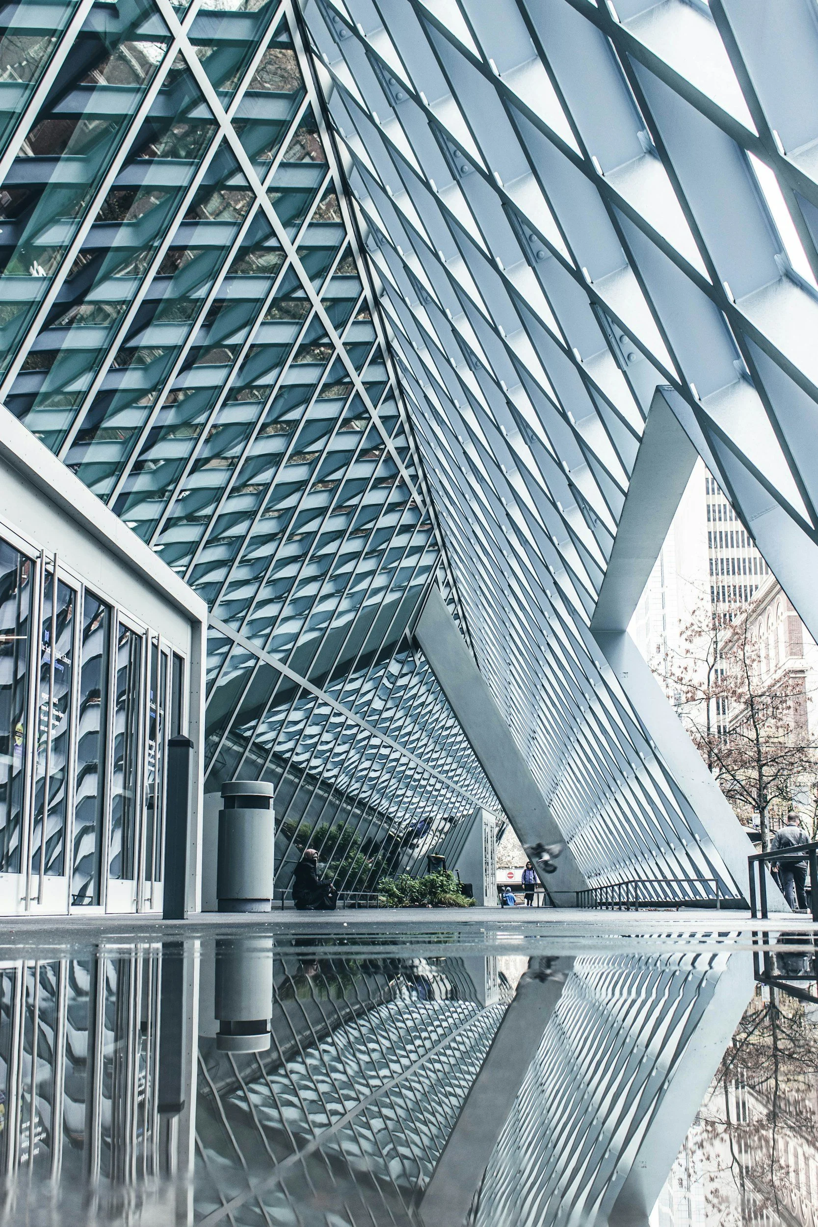Modern building with angled glass facade and metal framework, reflecting in a puddle on the sidewalk. A few people are seated and walking nearby.