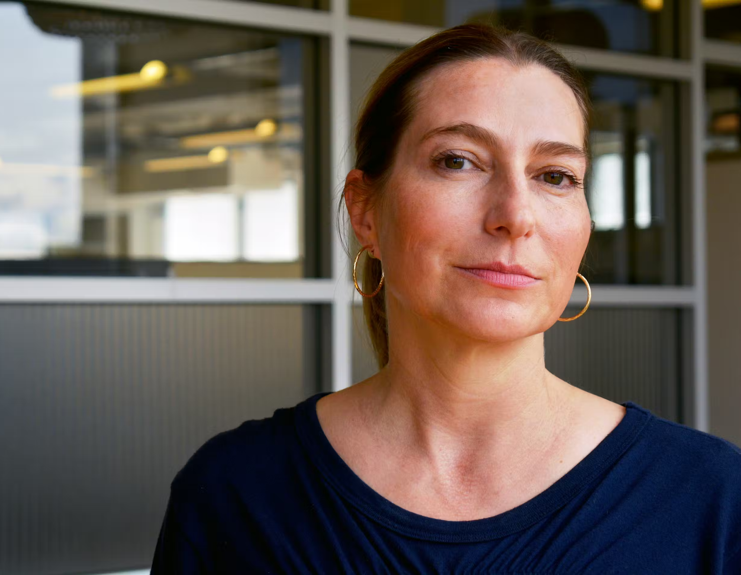 A woman with shoulder-length brown hair, wearing gold hoop earrings and a navy blue top, standing indoors in front of glass partition walls.
