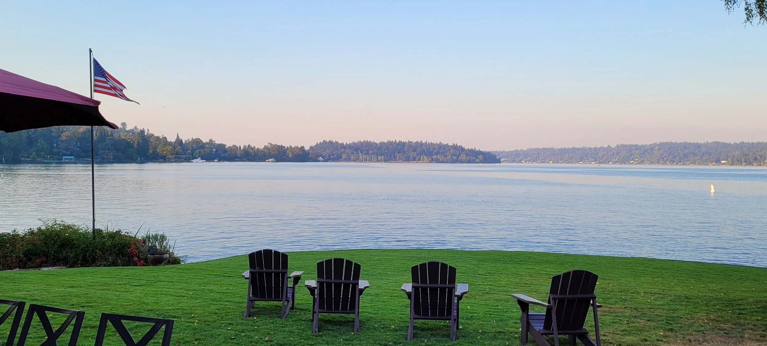 A lakeside view with four black Adirondack chairs facing the water, a green lawn, an American flag on a pole, a partially visible purple patio umbrella, and distant tree-covered hills across the lake.