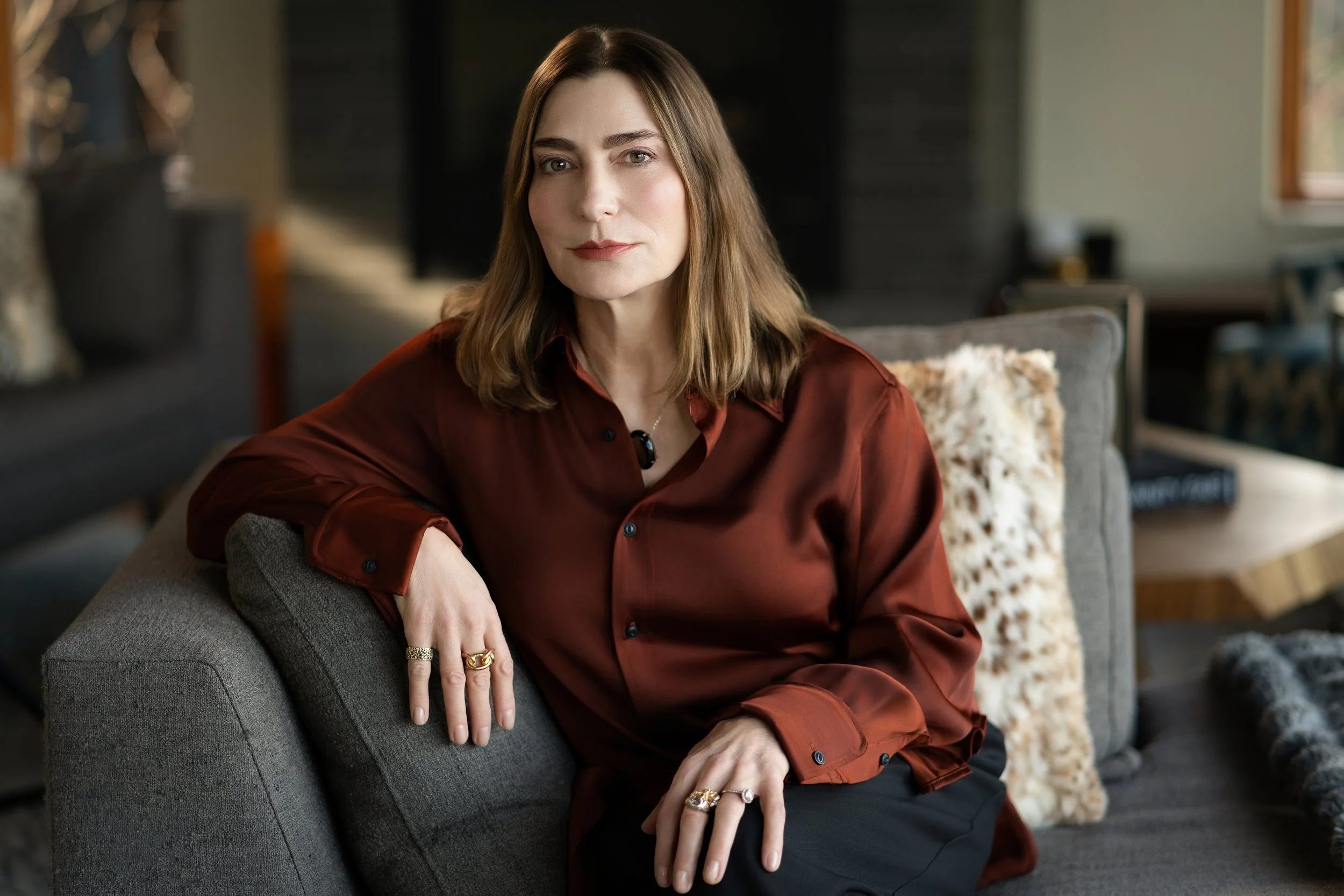 A woman with shoulder-length brown hair, wearing a satin burgundy blouse, sits on a gray sofa with a decorative pillow behind her, in a cozy living room.