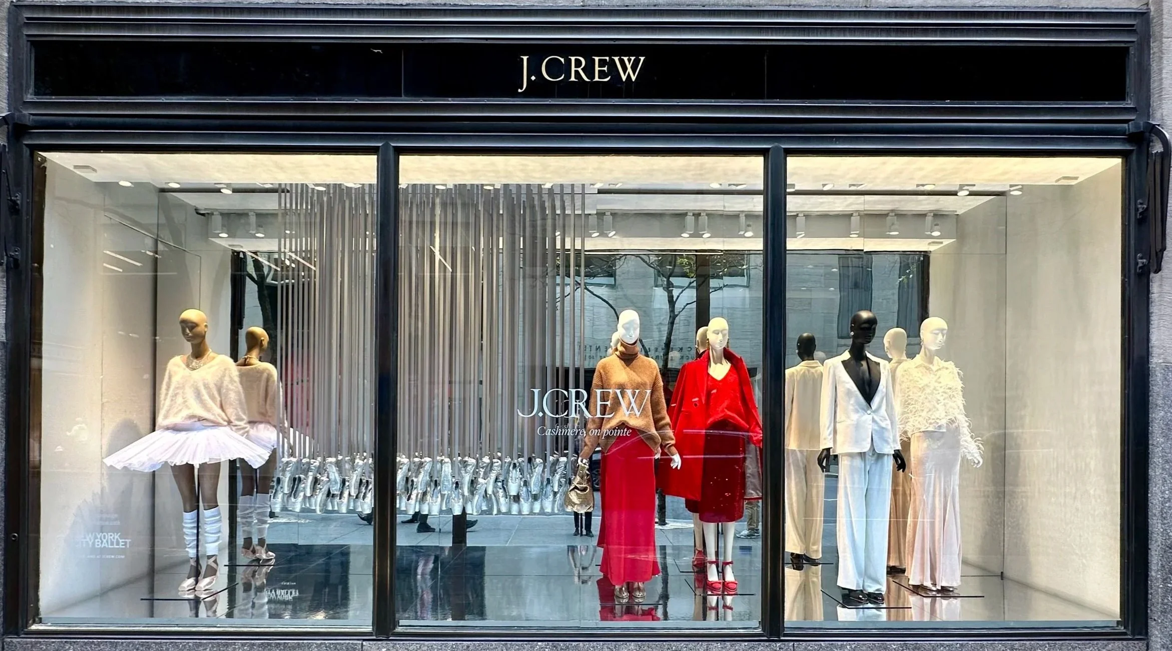 Fashion mannequins display elegant outfits in a store window, with a variety of clothing styles including dresses, suits, and outerwear, behind glass doors.