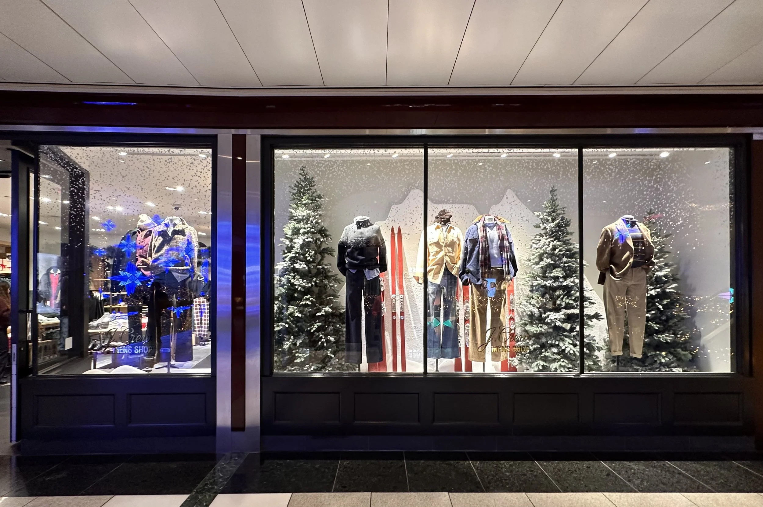 Store window display decorated for winter with three mannequins dressed in winter clothing, surrounded by snow-covered evergreen trees and a snowy backdrop, with a mountain sketch in the background.