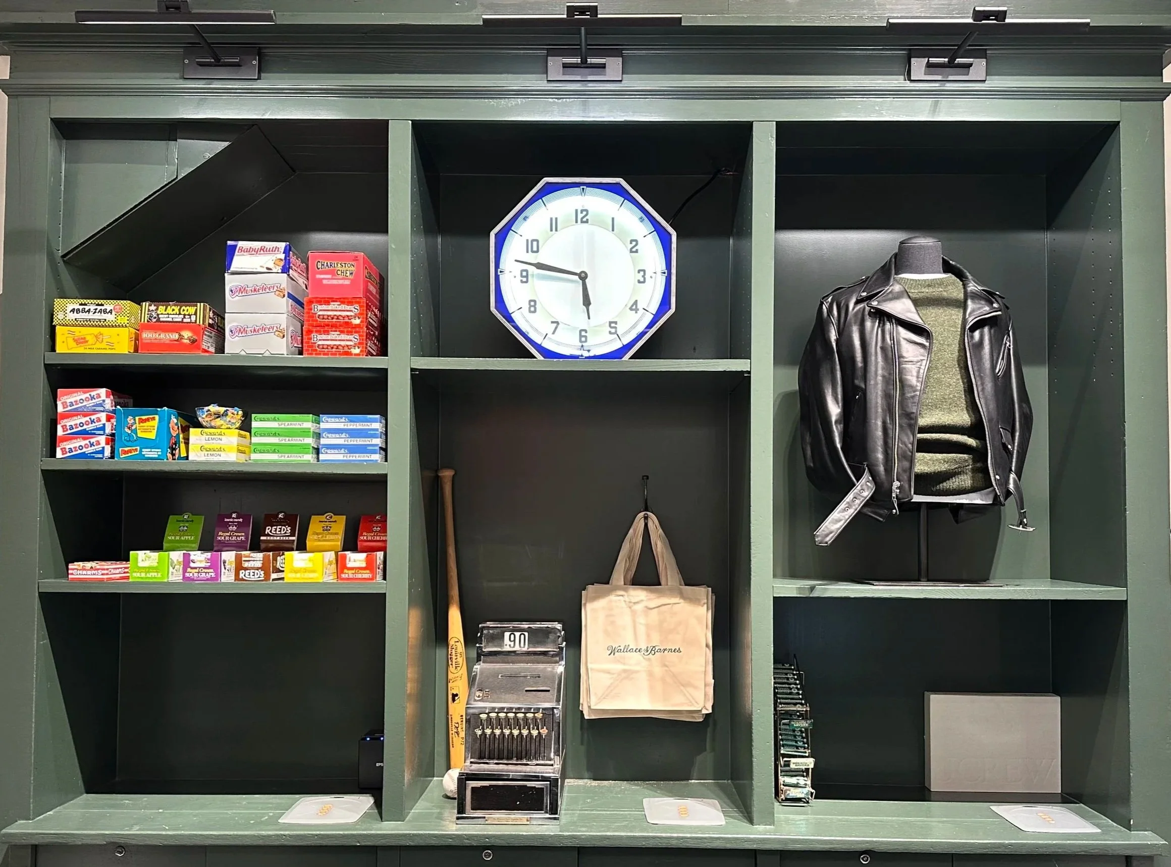 Green display shelf with snacks, a clock, a baseball bat, a tote bag, a black leather jacket on a mannequin, and a cash register.