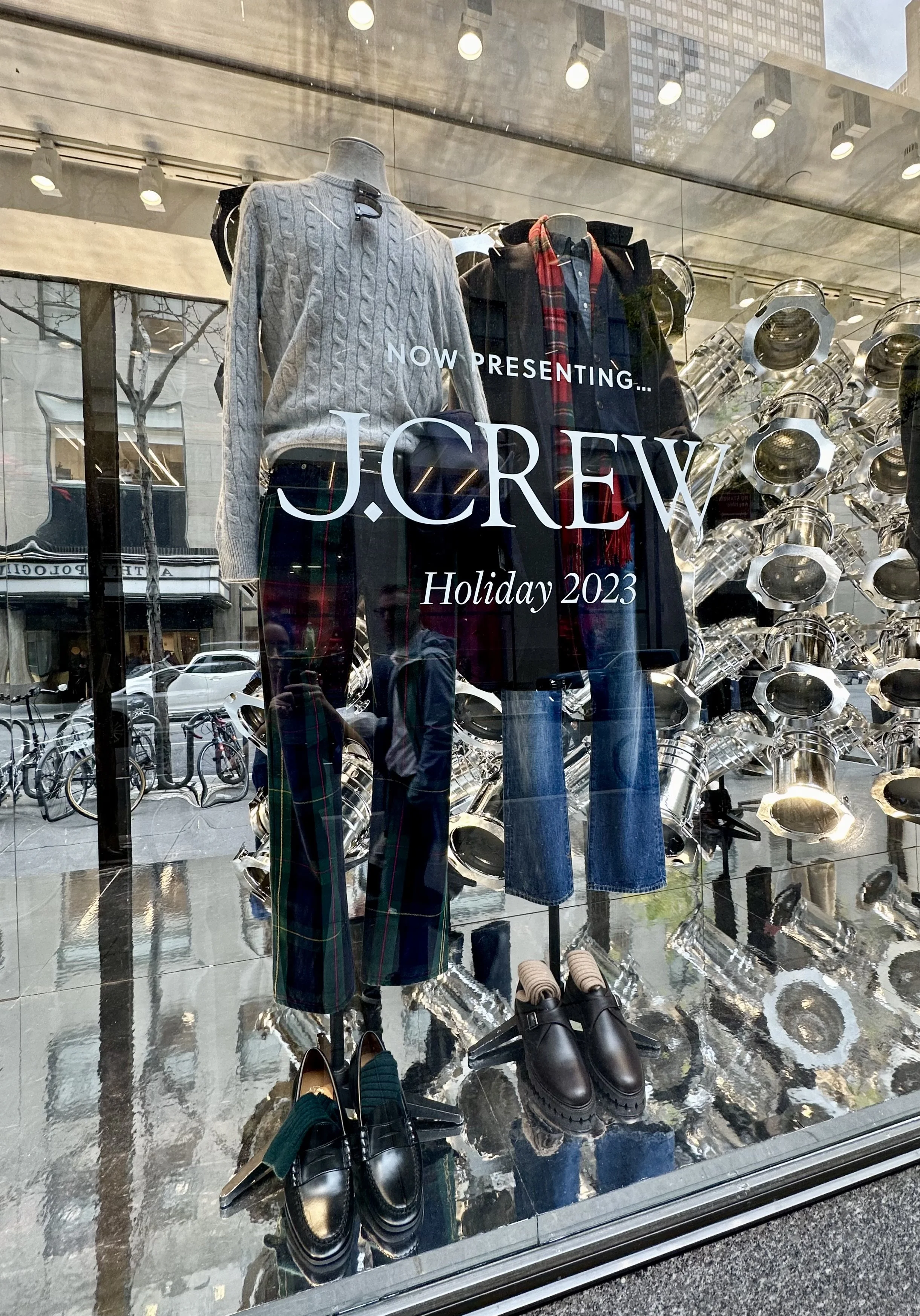 J.Crew store window display featuring mannequins dressed in fall and winter clothing, including sweaters, plaid pants, coats, and boots, with a background of decorative metallic objects and street view reflections.