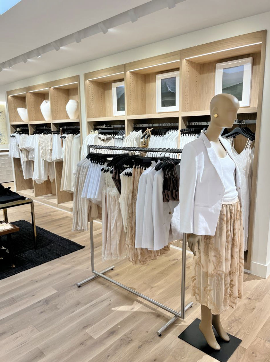 Clothing store display featuring white and beige garments on mannequins and racks, with wooden shelving units and small windows in the background.