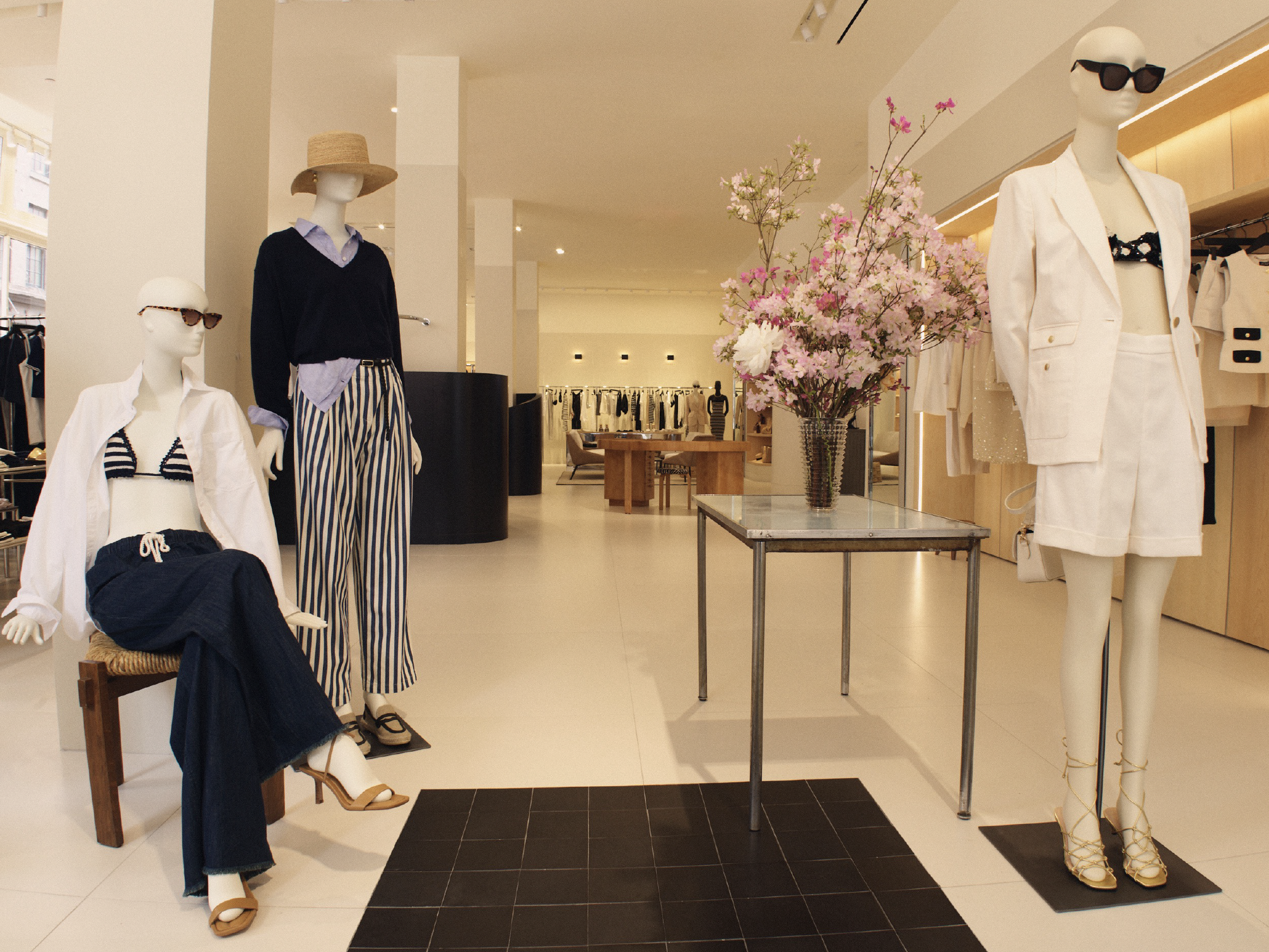Display of mannequins dressed in fashionable summer outfits in a clothing store, with a table holding a large vase of pink flowers nearby.