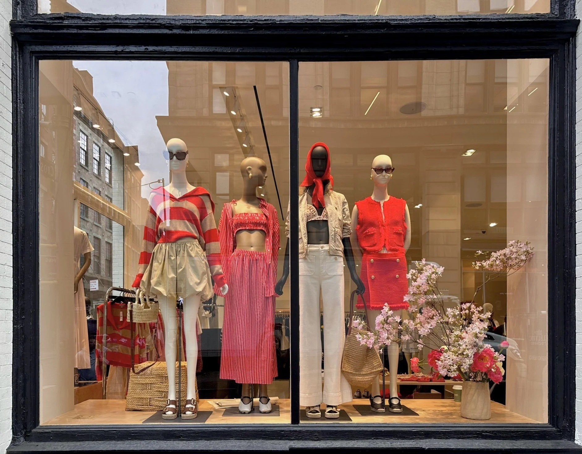 Mannequin display in a clothing store window featuring five mannequins dressed in red and white summer outfits, with pink flowers in a vase in front.