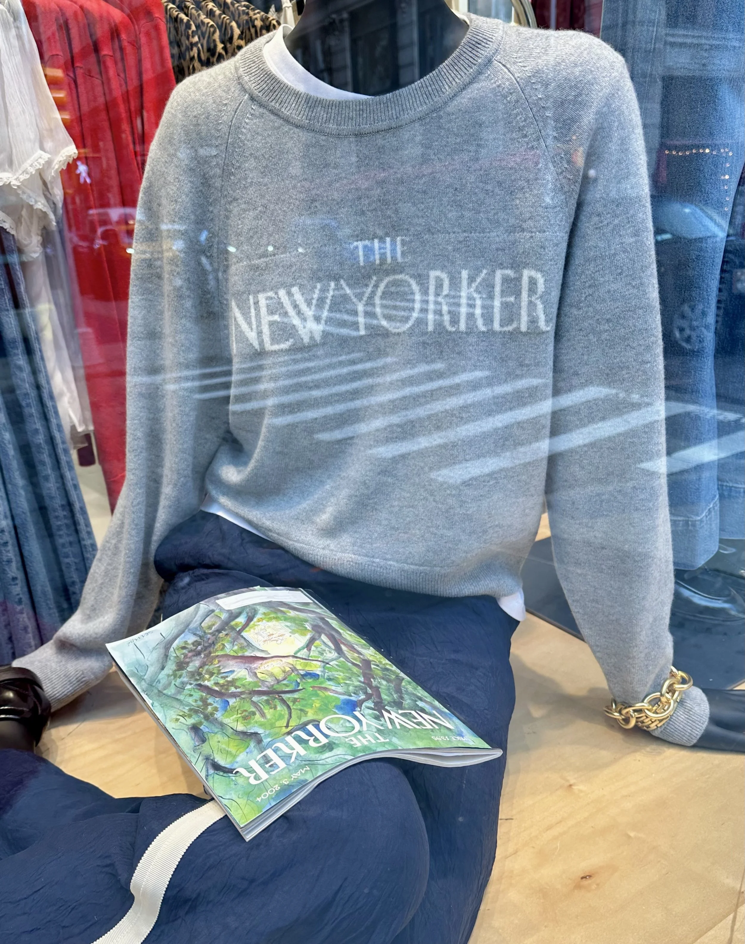 Mannequin wearing a grey sweatshirt with 'The New Yorker' logo in a retail store window, holding a colorful magazine titled 'The New Yorker'.