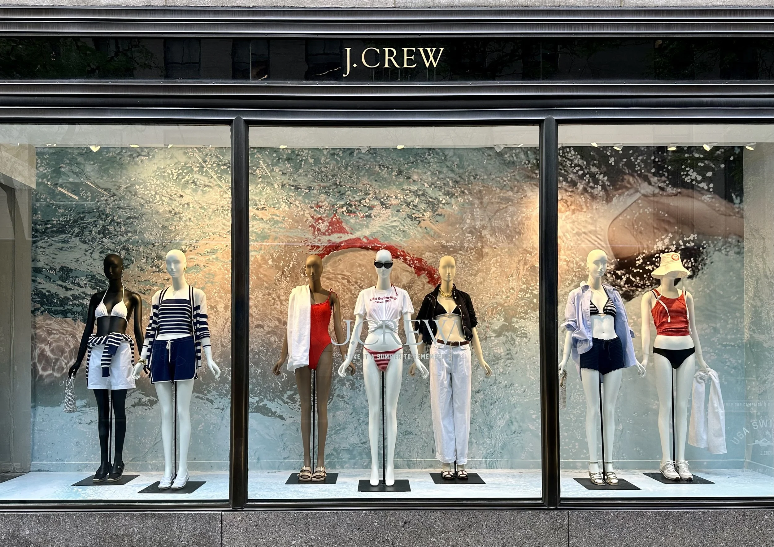 Storefront window display with mannequins dressed in summer swimwear and casual clothing, with an ocean wave background and the brand name J.Crew at the top.