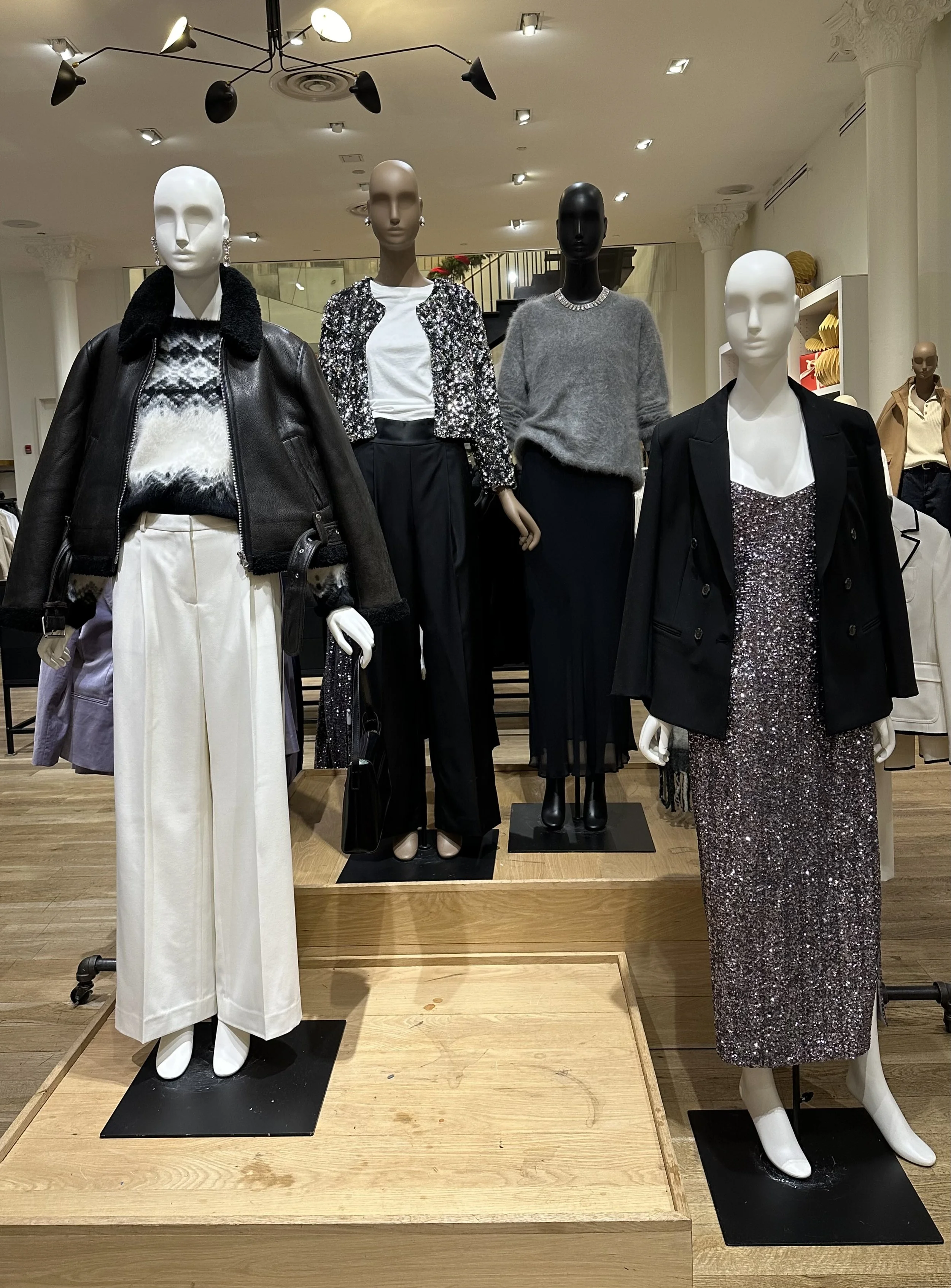 Display of five mannequins showcasing fashionable women's clothing, including a black leather jacket with a fur collar, a sequin jacket, a gray sweater, and a sparkly dress, in a retail store.