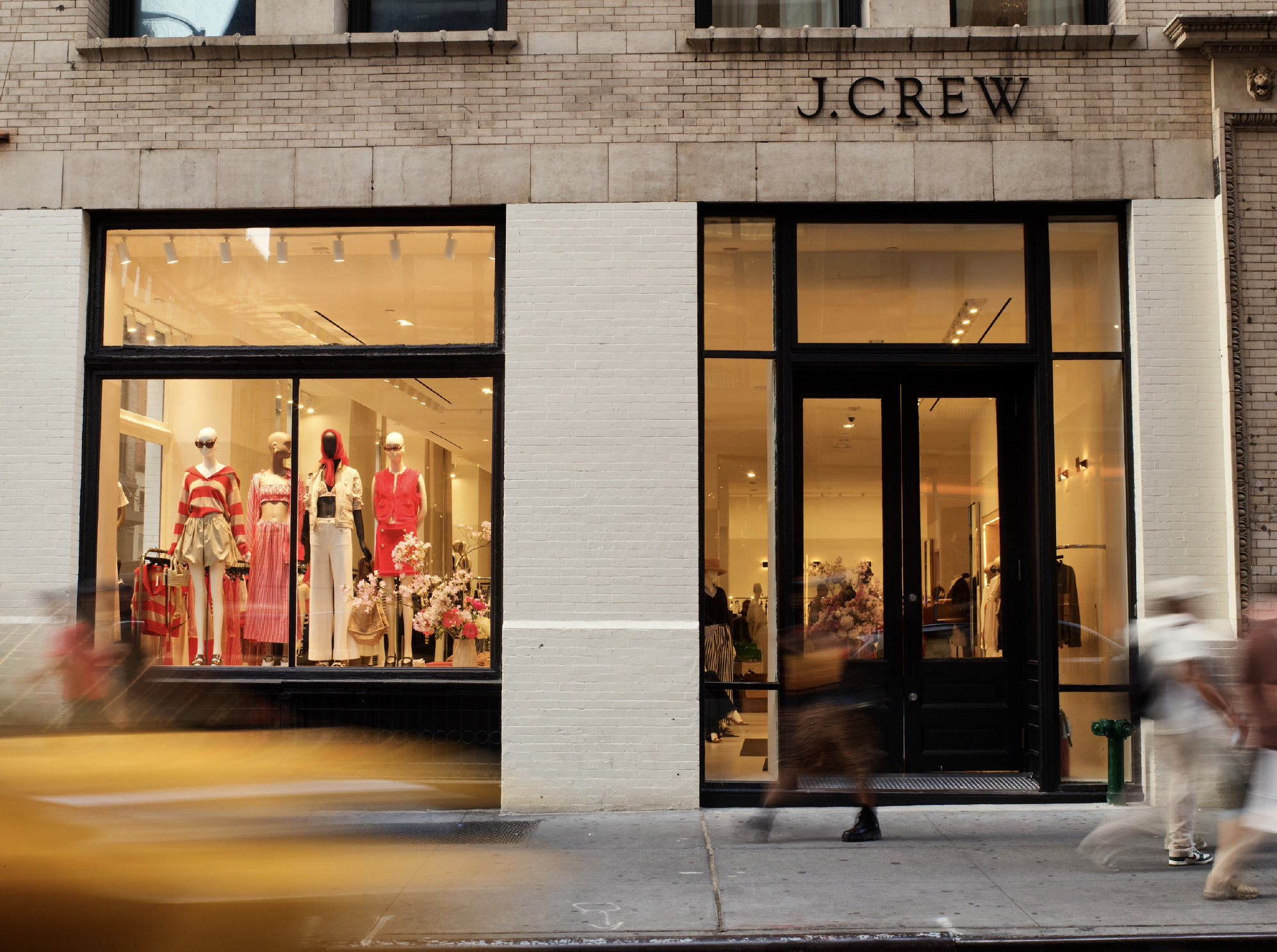 Storefront of J.Crew with display window showing mannequins dressed in colorful clothing and flowers, black door and large windows, pedestrians walking by.