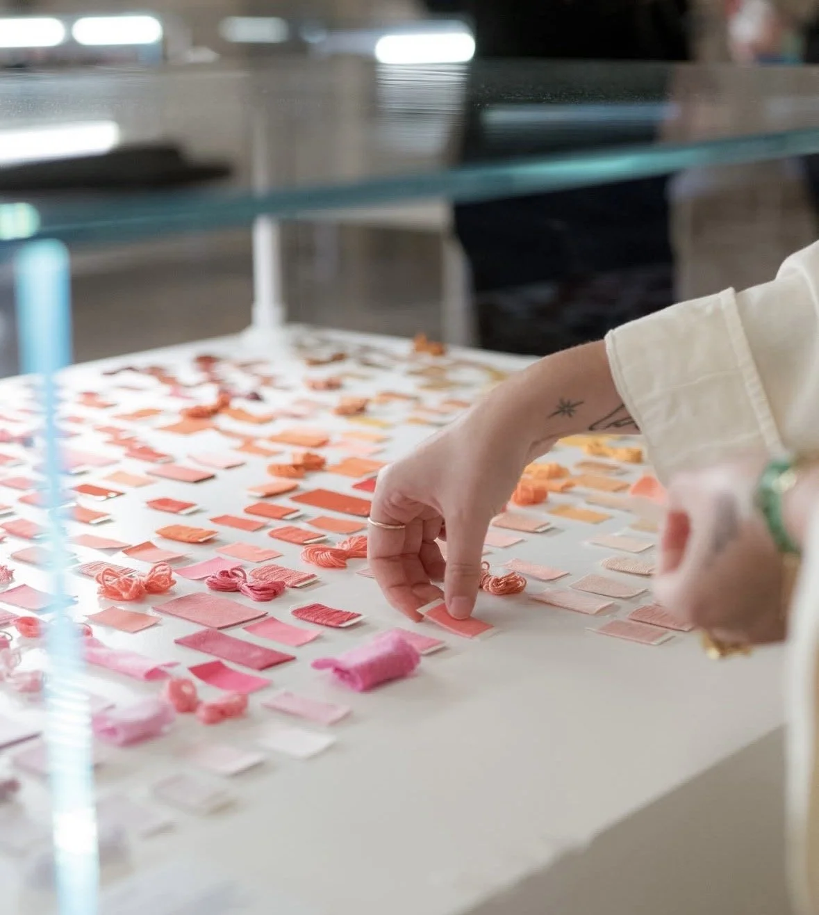 A person with a tattoo on their arm sorting through small fabric swatches in shades of pink, peach, and orange on a white table.