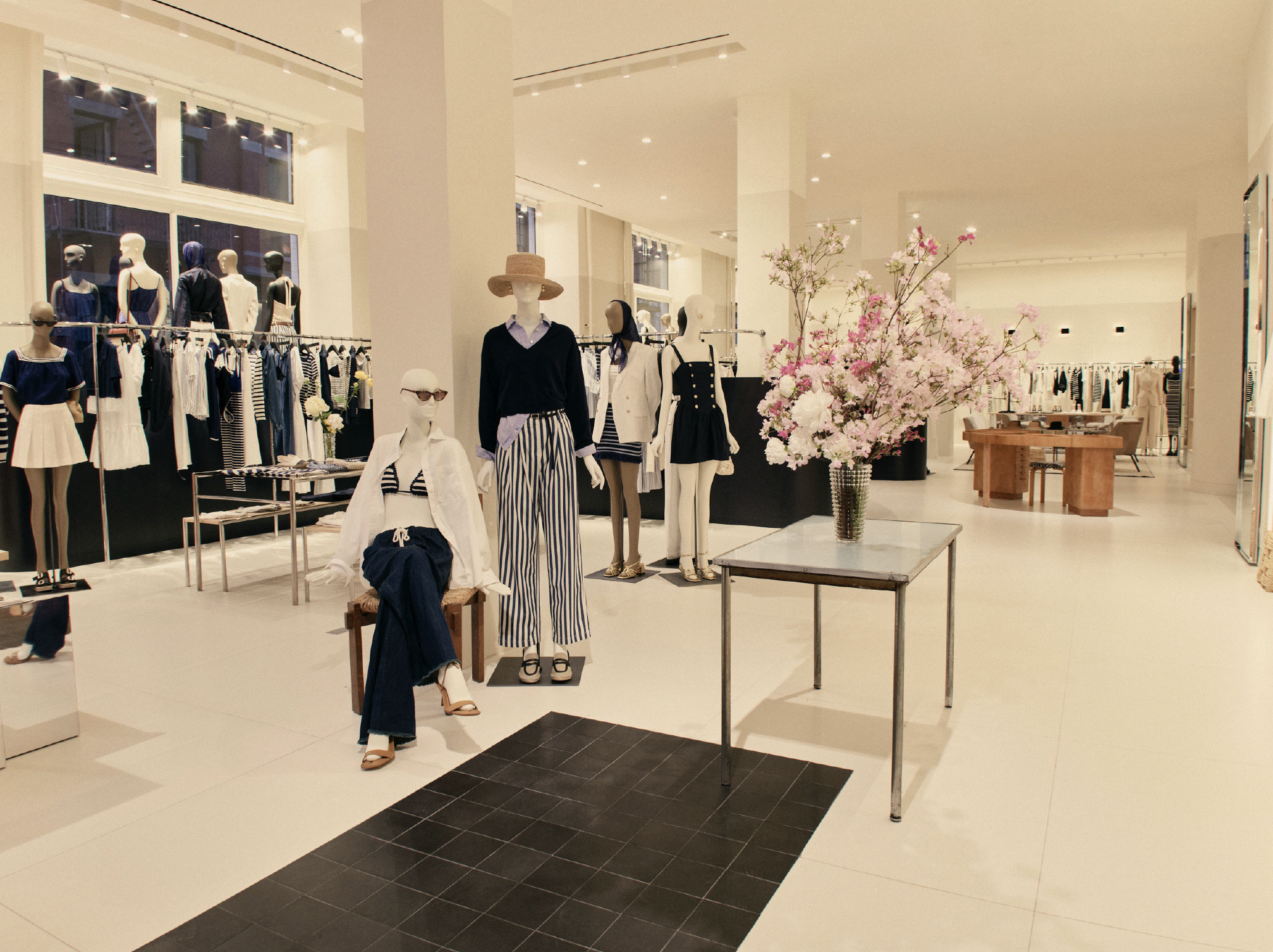 A retail clothing store with mannequins displaying summer outfits. The store has white walls and high ceilings, with large windows letting in natural light. There is a large floral arrangement of pink and white flowers on a glass table near the mannequins.