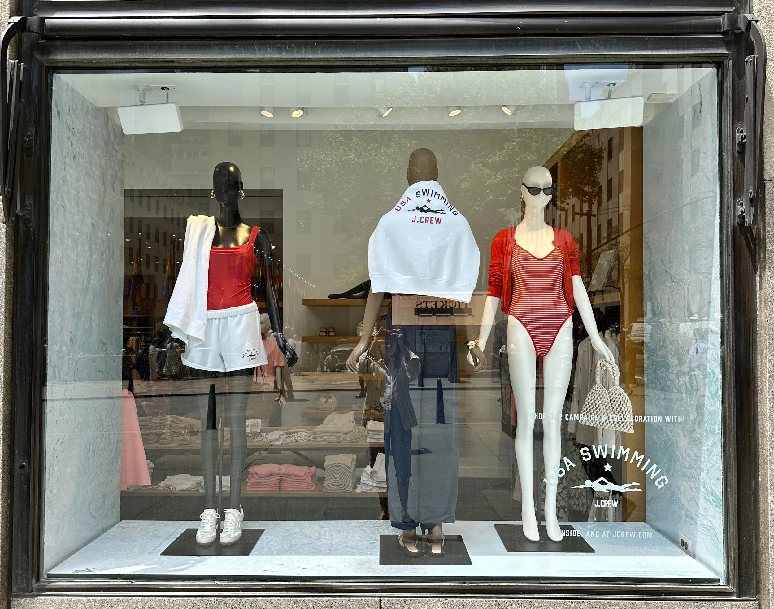 Store window display with mannequins dressed in swimwear and casual clothing, featuring red and white outfits, along with the brand logo 'J.Crew' and promotional text.