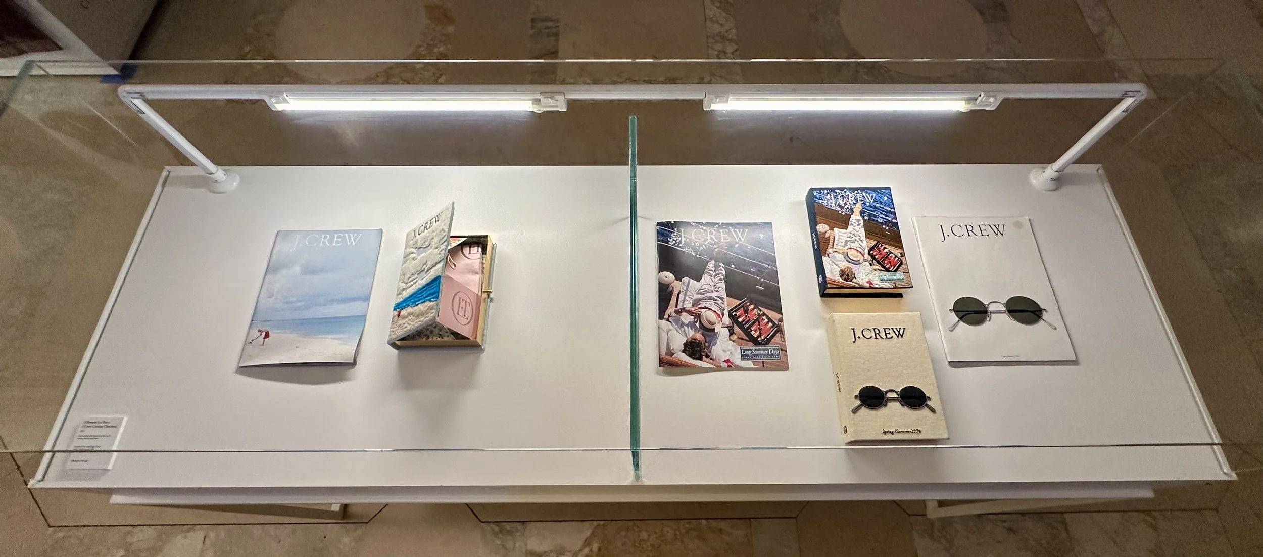 Display table with open magazines, books, and sunglasses from J.Crew, arranged symmetrically, with magazine covers featuring beach scenes, summer accessories, and models.