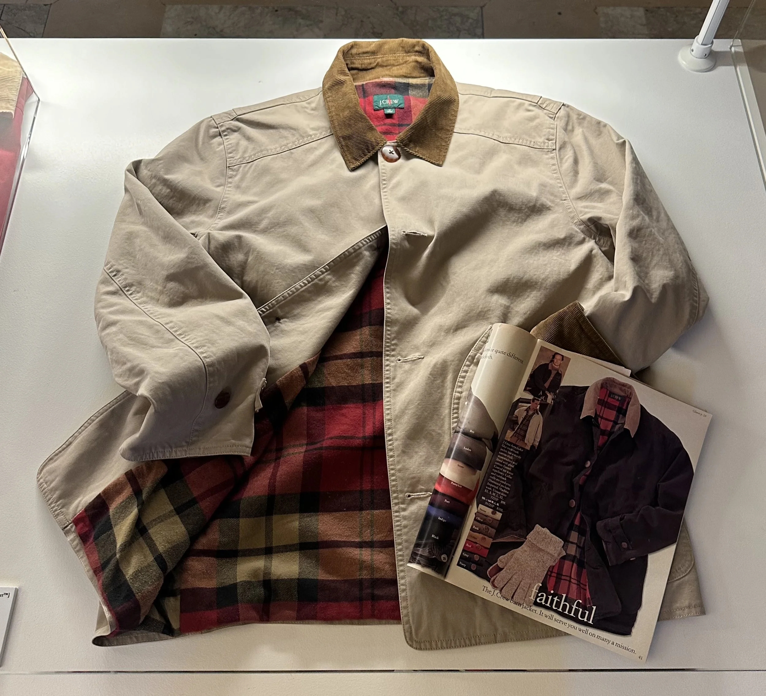 A beige jacket with a tan corduroy collar layered over a red and black plaid shirt, opened to show the shirt's collar and part of its pattern, with an open magazine featuring a similar jacket and shirt combination.