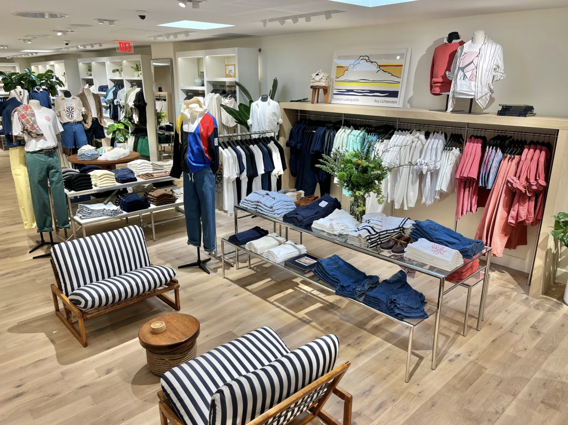 Clothing store interior with mannequins, racks of shirts, pants, and folded clothes, and a seating area with striped cushions and a small wooden table.