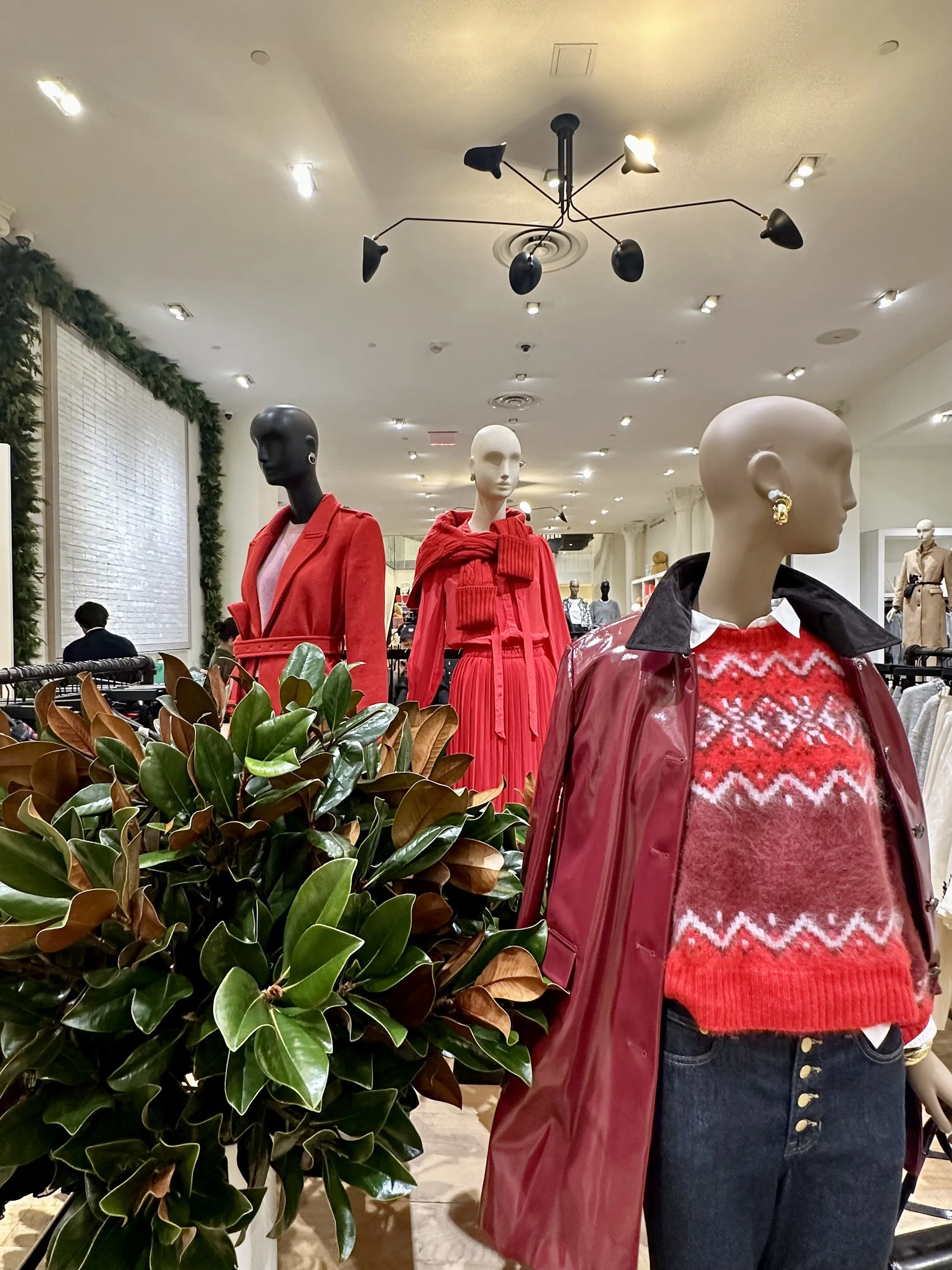 Mannequins in a clothing store display dressed in red outfits, with green plants in the foreground and store interior visible in the background.