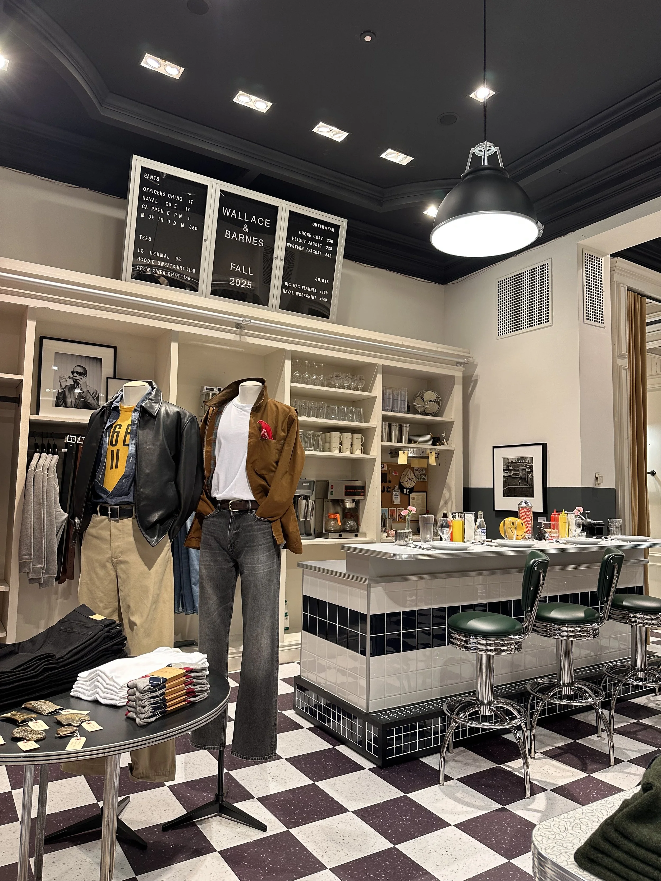 Interior of a stylish cafe or diner with mannequins displaying casual clothing, a checkered floor, a bar counter with green stools, and shelves with glasses and cups. There are framed photos, a coffee machine, and a bulletin board.
