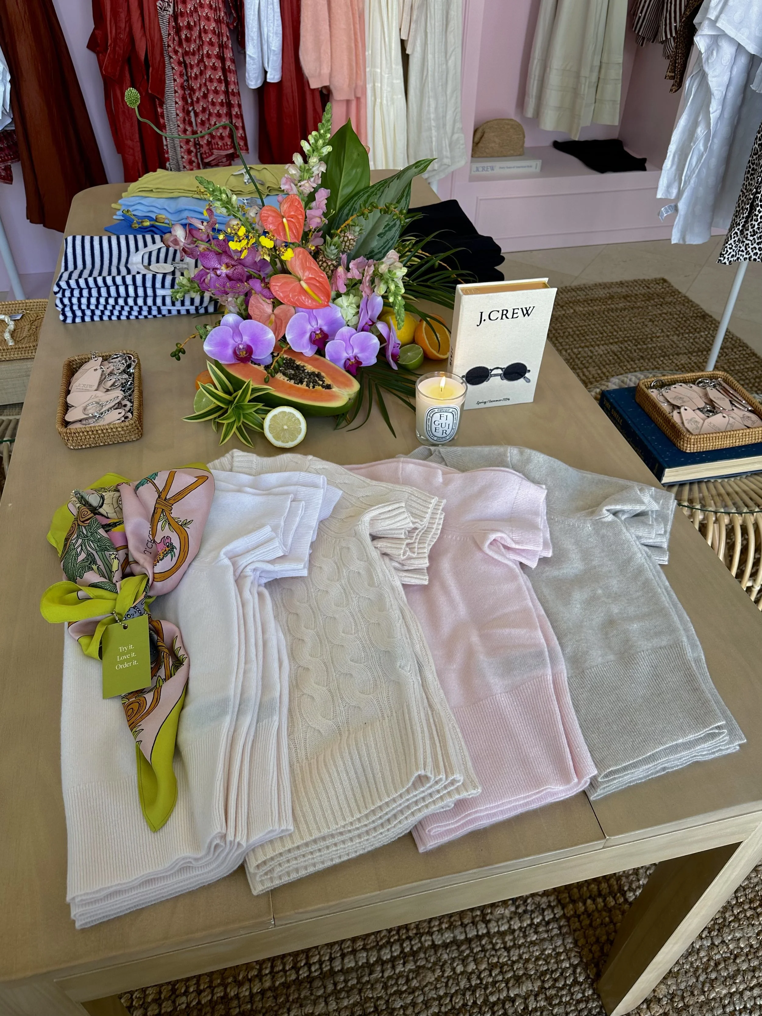 Array of folded pastel sweaters, a silk scarf, and accessories in a boutique display with a floral arrangement, sliced fruit, a candle, and a fashion catalog on a wooden table.