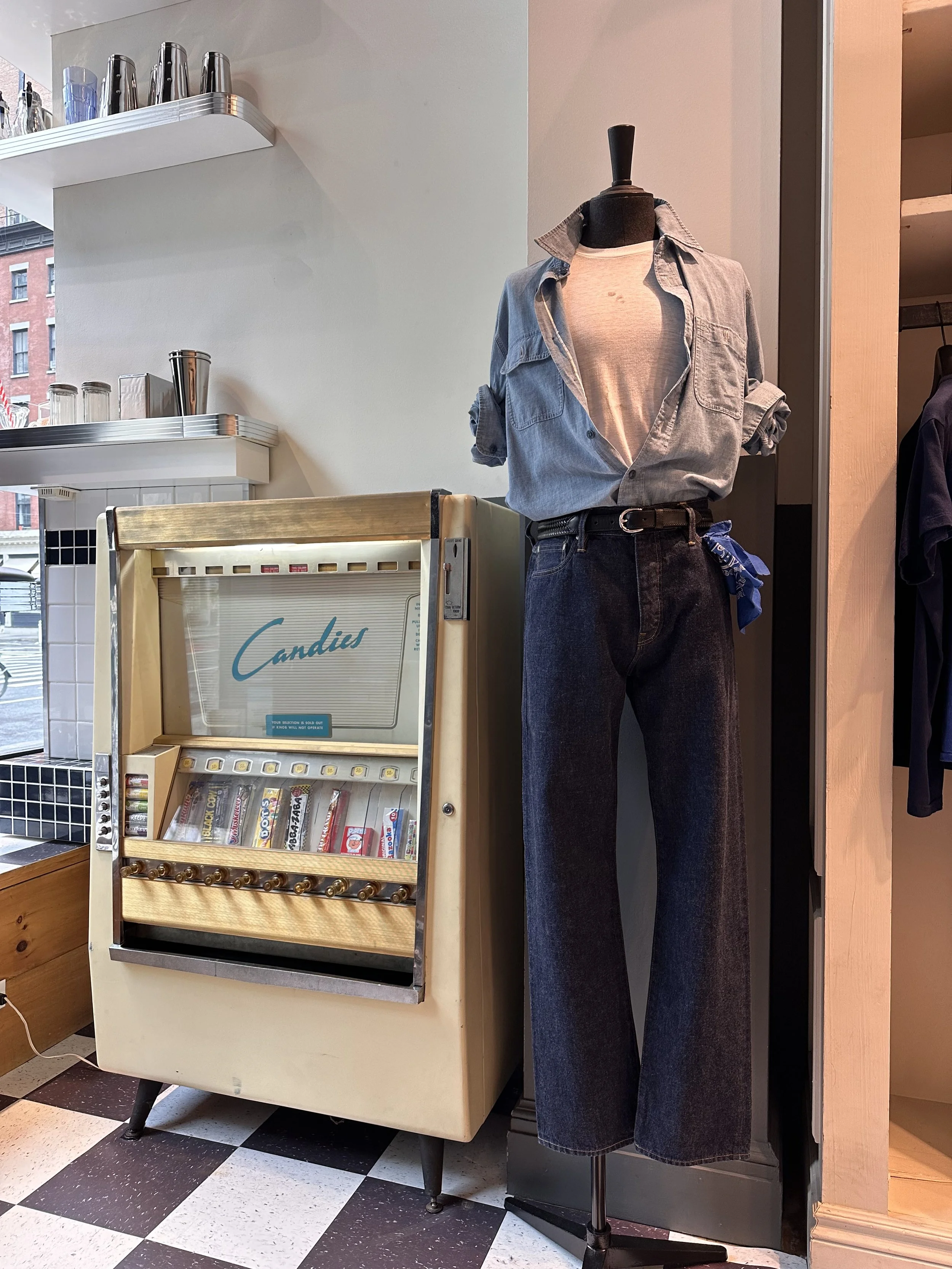 A mannequin dressed in a light gray denim button-up shirt with rolled-up sleeves, a white T-shirt underneath, and high-waisted dark jeans with a blue bandana tied to the belt loop, standing next to a vintage vending machine with candies in a cafe or shop.
