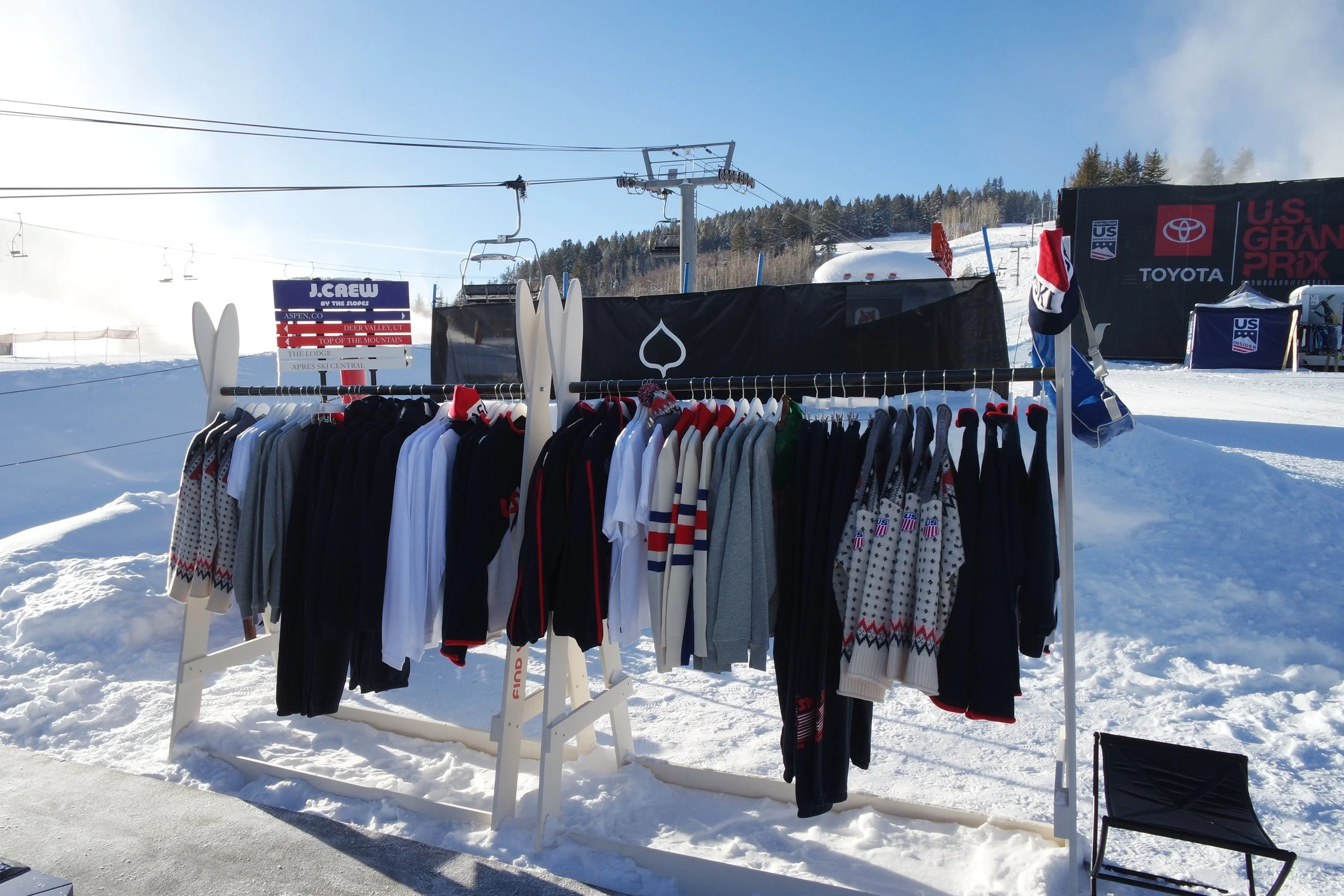 Clothes displayed on a rack outdoors on snow at a ski resort, with signs and ski lifts in the background.