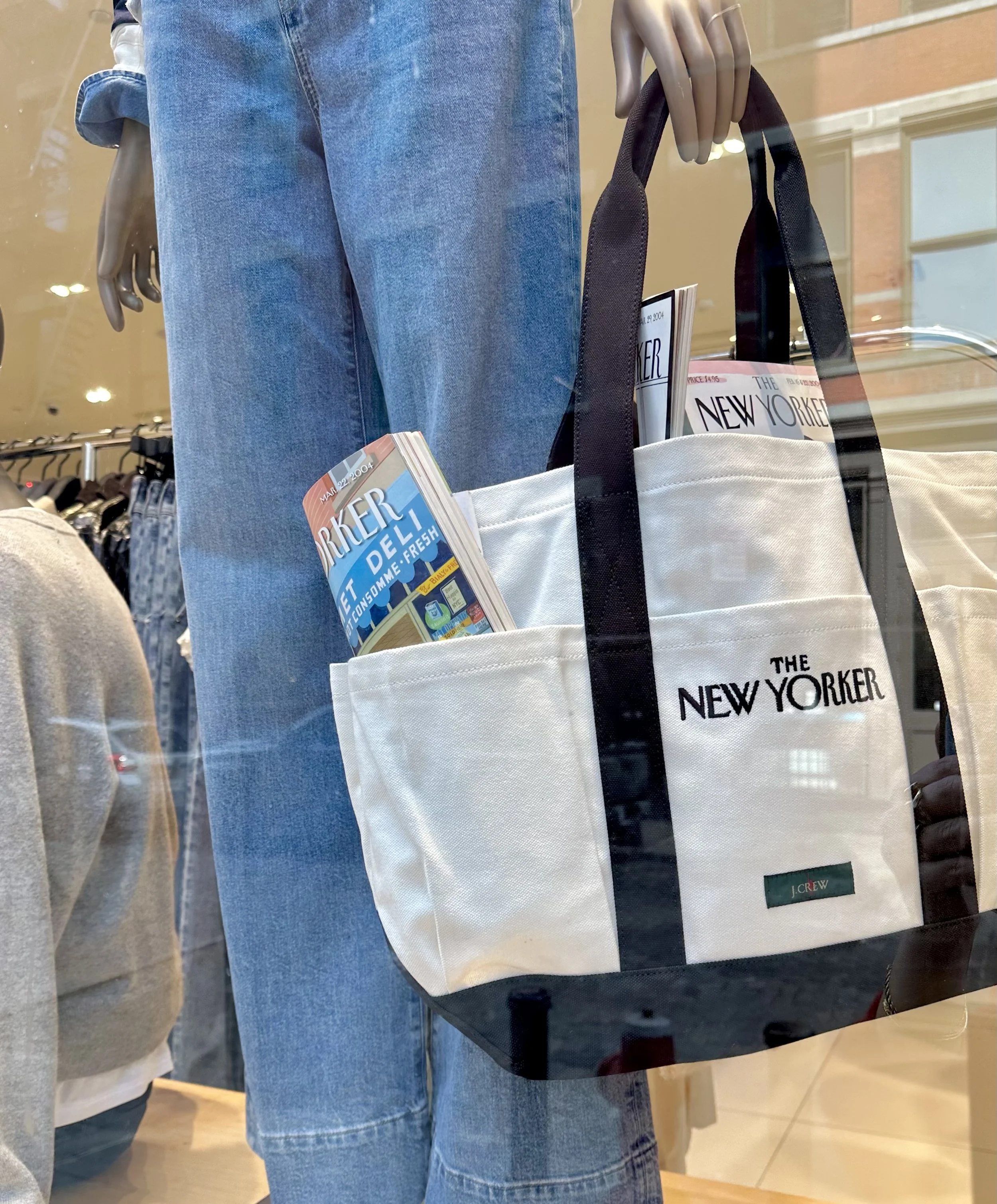 A retail store window display featuring a mannequin wearing blue jeans, holding a white and black tote bag with the words 'The New Yorker' printed on it, containing magazines and pamphlets, including one titled 'The New Yorker'.