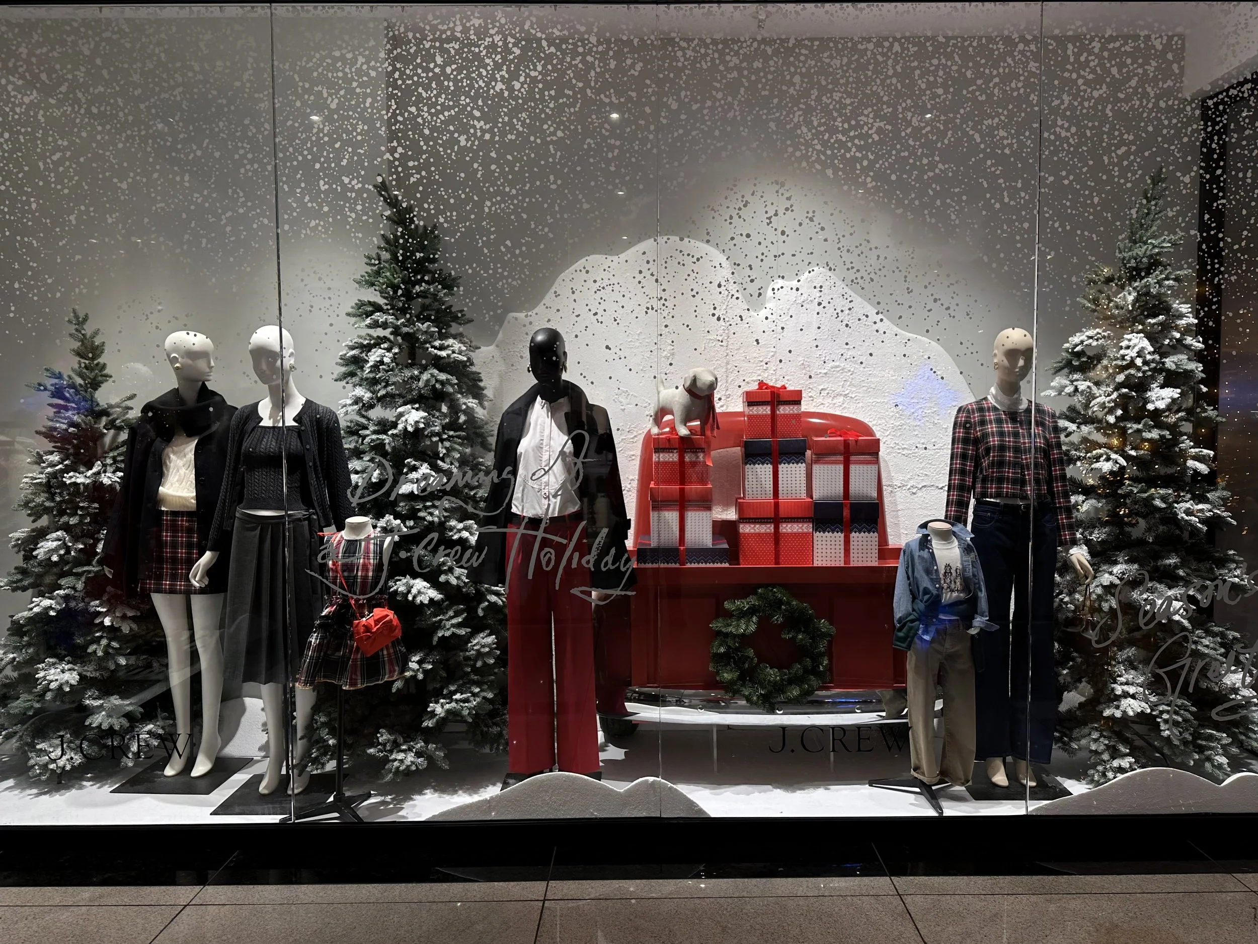 Holiday-themed clothing display in store window with mannequins dressed in winter outfits, snow-covered trees, and a large red sled filled with wrapped Christmas presents, with a white textured backdrop resembling snow.