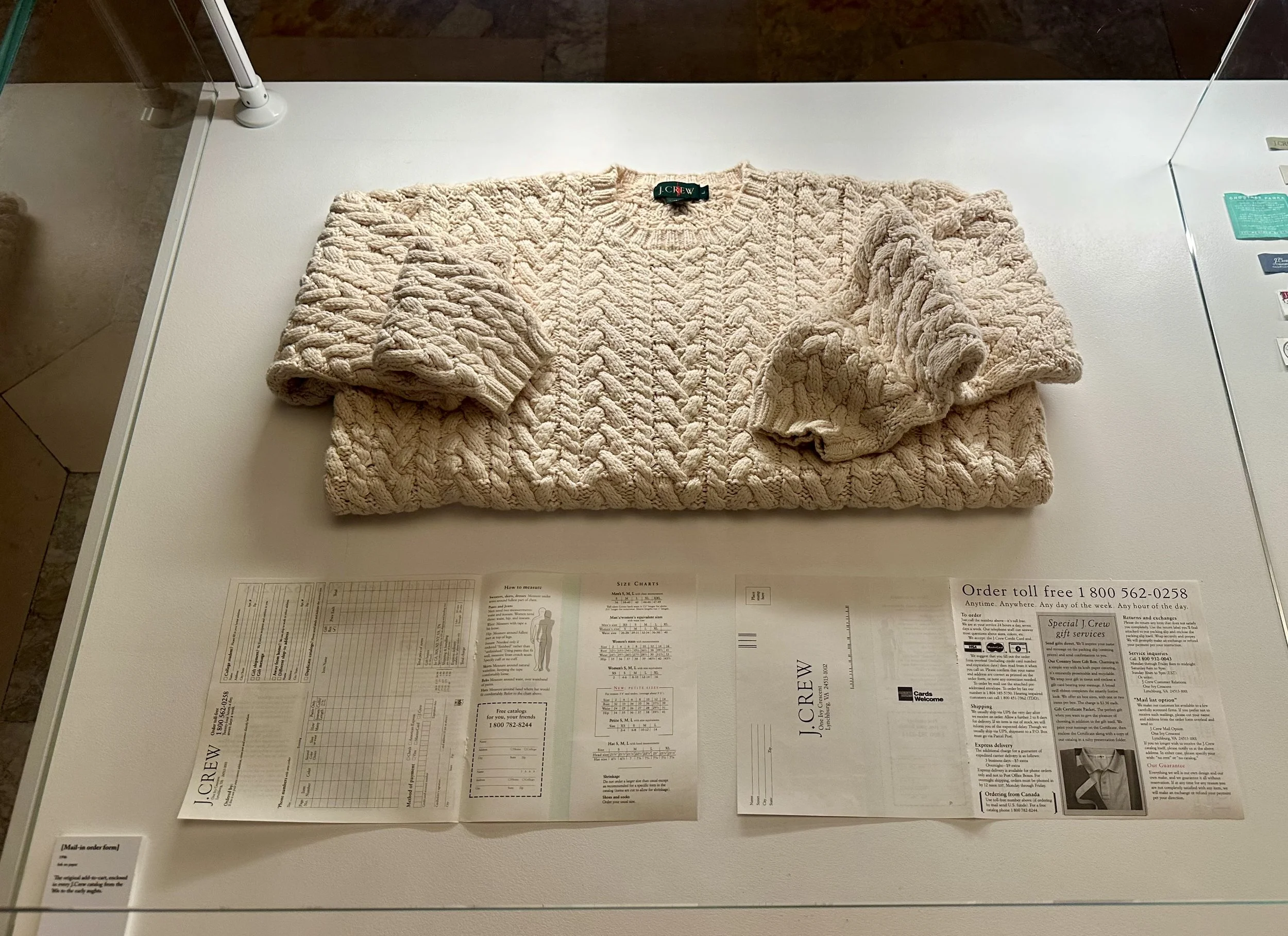 A beige chunky knit sweater with long sleeves laid flat on a white display table in a glass case, with printed material including a sales sheet or catalog underneath.