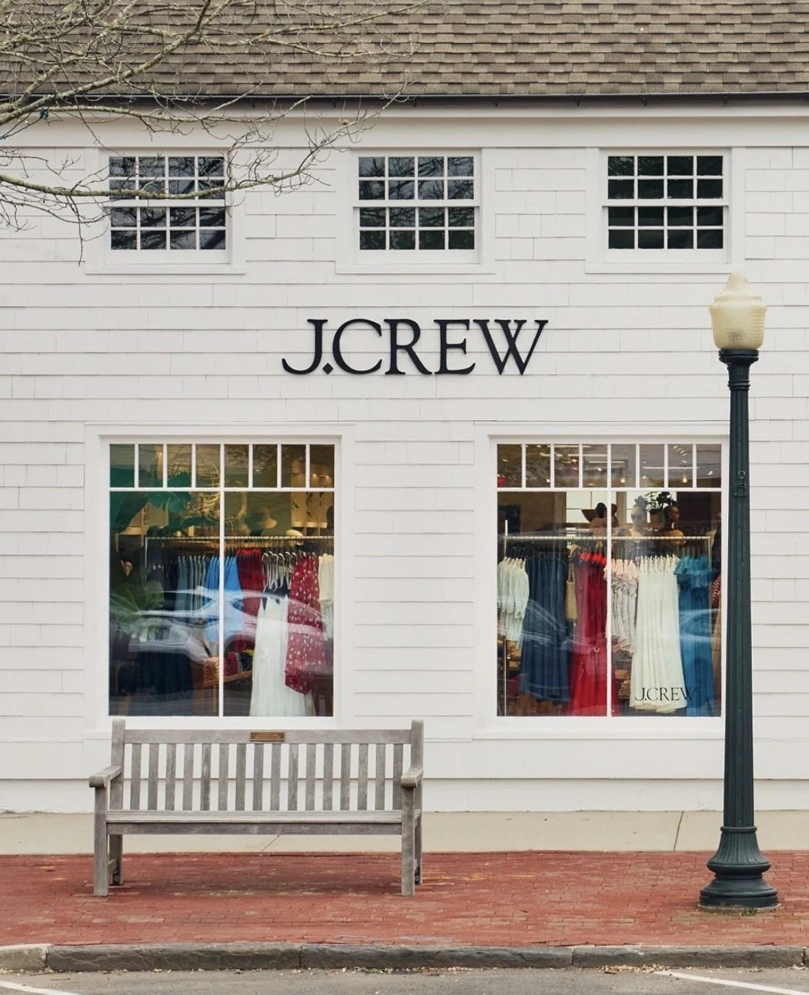 The storefront of J.Crew with large windows displaying dresses and a bench outside.