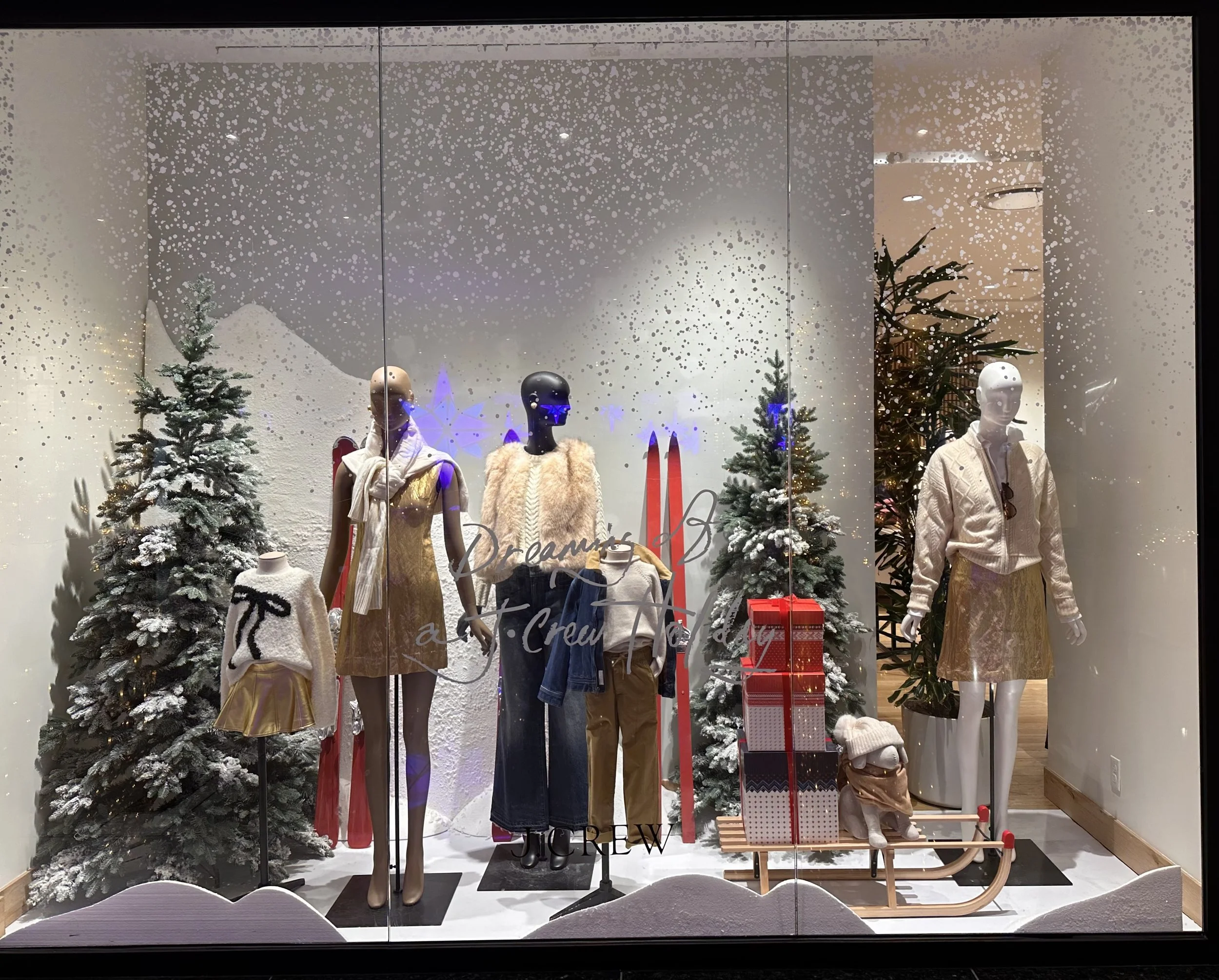 Mannequins in a holiday display window with snow-covered Christmas trees, wrapped presents, and winter clothing, including dresses, sweaters, and jackets, featuring gold and neutral tones.