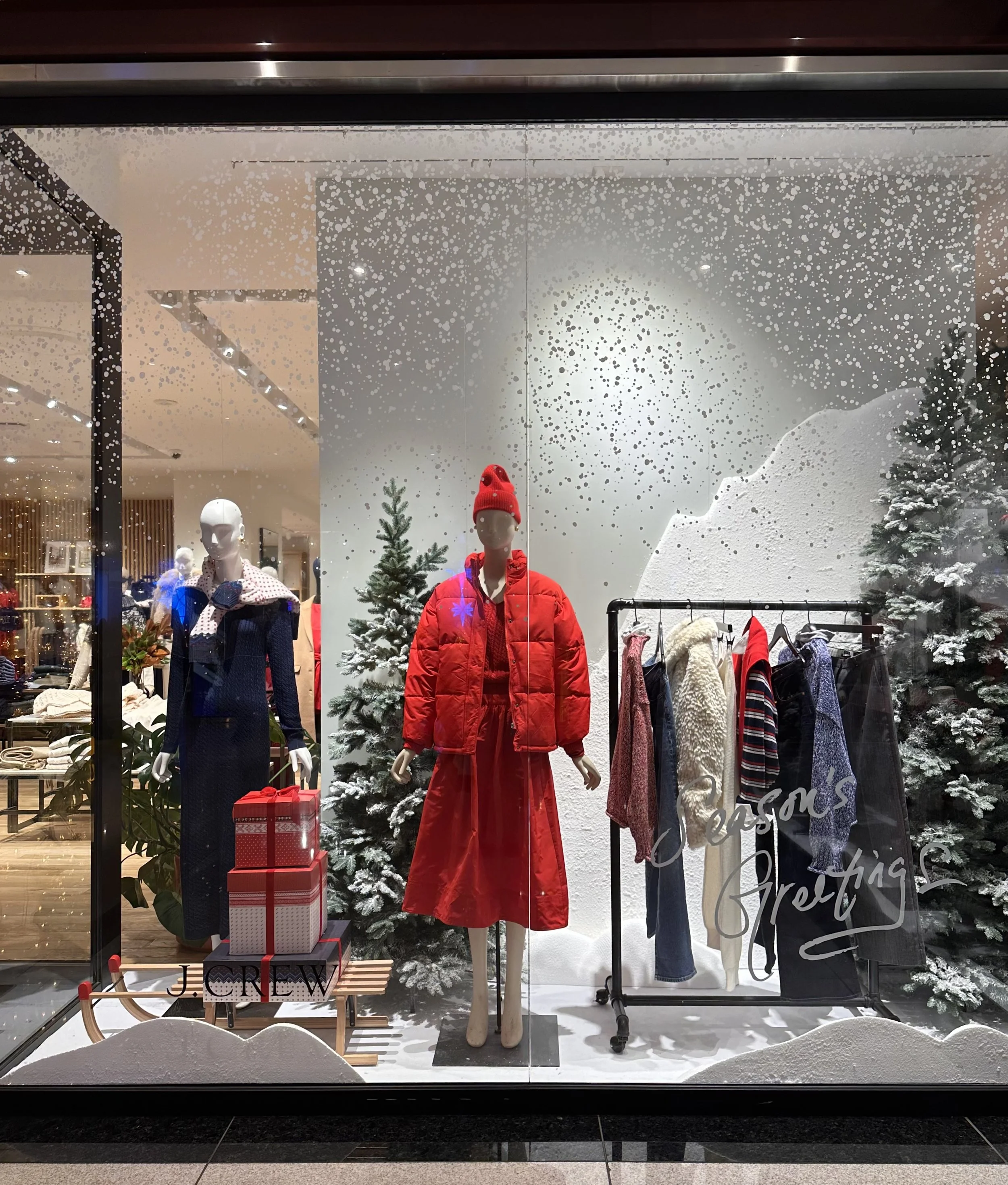 Store window display with mannequins dressed in winter clothing, including a red jacket, red skirt, and red beanie, surrounded by Christmas trees and holiday decor, with seasonal gift boxes and a "Season's Greetings" sign.