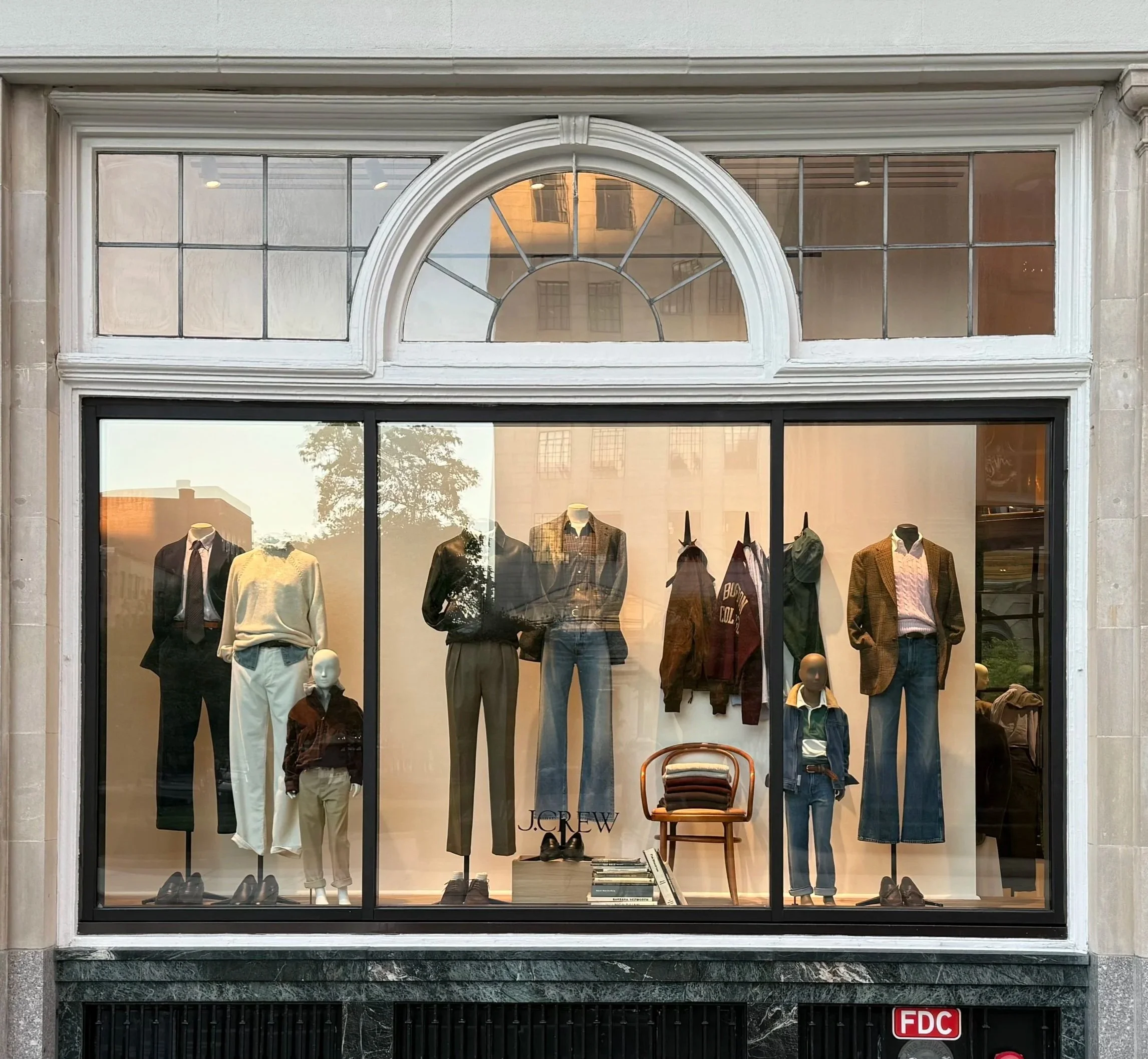 Storefront window display with mannequins dressed in men's fashion, including suits, jackets, and casual clothes, with a chair and books in the center.