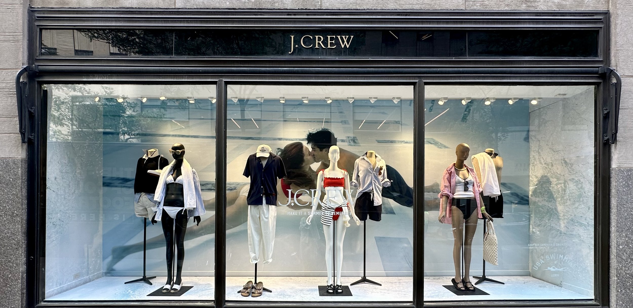 Display window of J.Crew boutique showcasing mannequins with casual summer outfits including swimsuits, shorts, and tops.