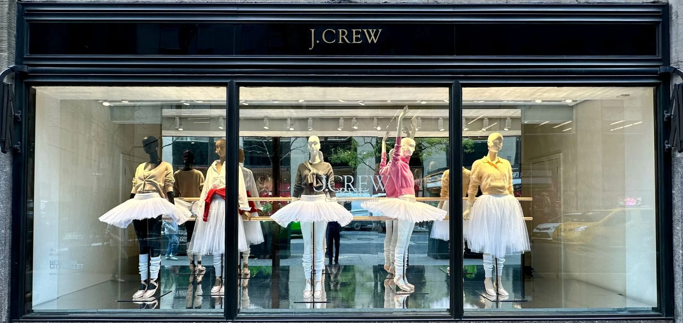 Display window of J. Crew store showcasing mannequins dressed in ballet-inspired outfits with tutus and casual tops.