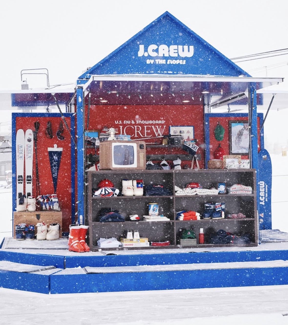 A ski and snowboard rental shop with J.Crew branding, set against snowy weather with falling snowflakes, displaying skis, boots, helmets, and clothing for winter sports for sale or rent.