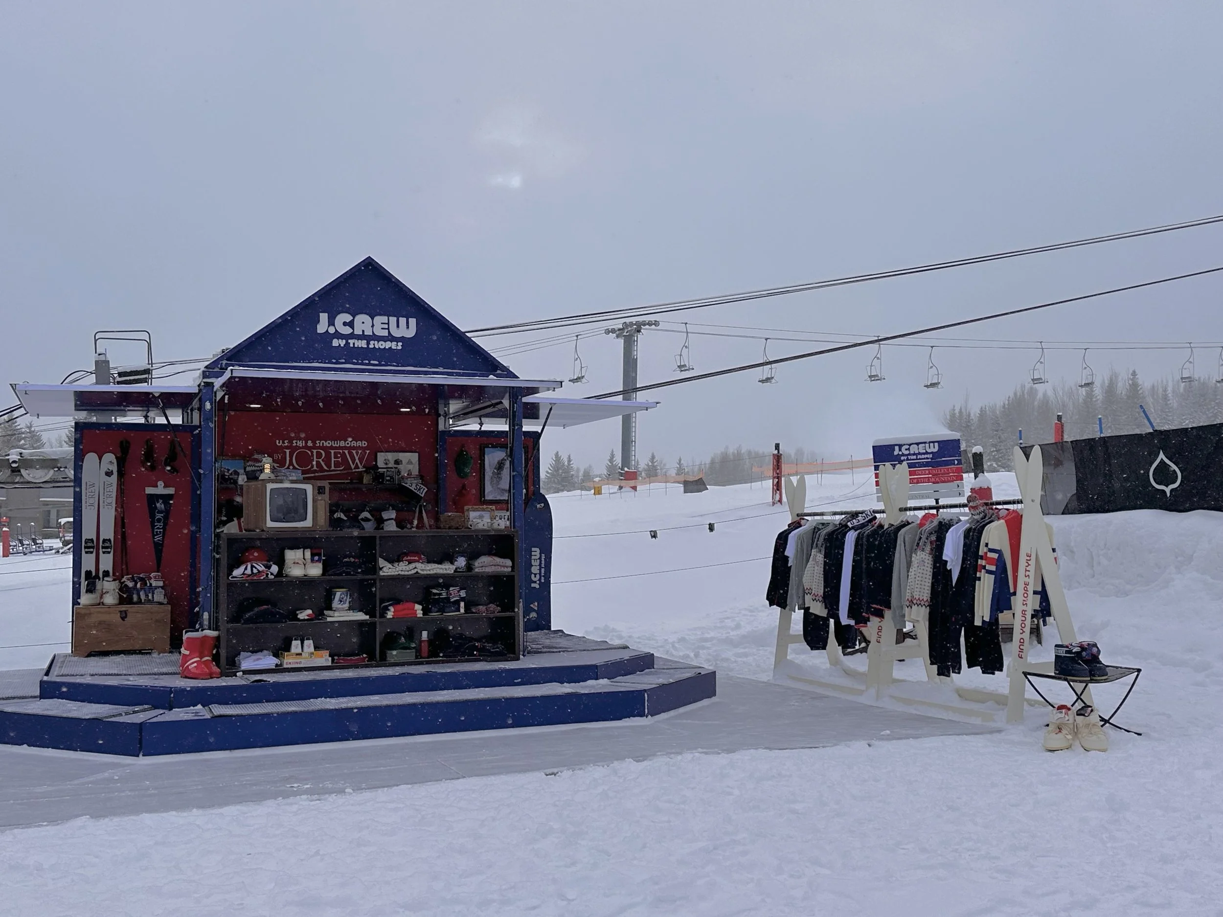 Snow-covered ski shop and clothing stand with ski lifts in the background at a ski resort.