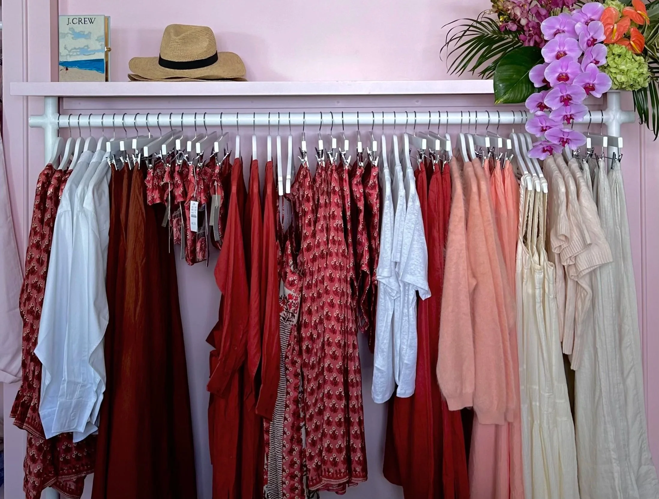 Clothing rack with various dresses and garments in red, white, peach, and beige on white hangers. Top shelf has a straw hat, a small framed picture, and a bouquet of large, colorful flowers with purple orchids. The background wall is pink.