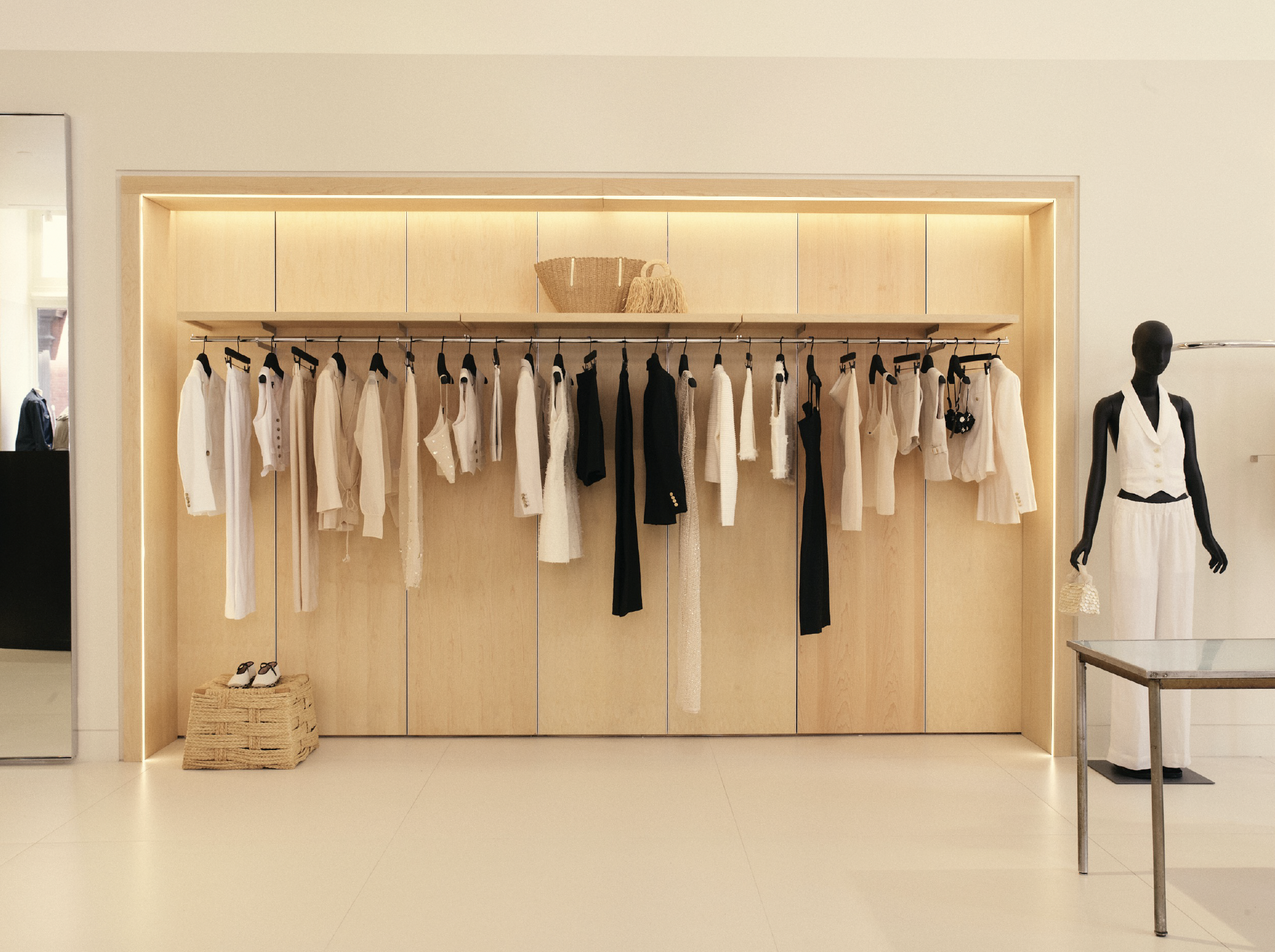 Clothing display in a boutique with white and black garments hanging on a rod, a wicker basket on top, and a mannequin dressed in white pants and a sleeveless top on the right.