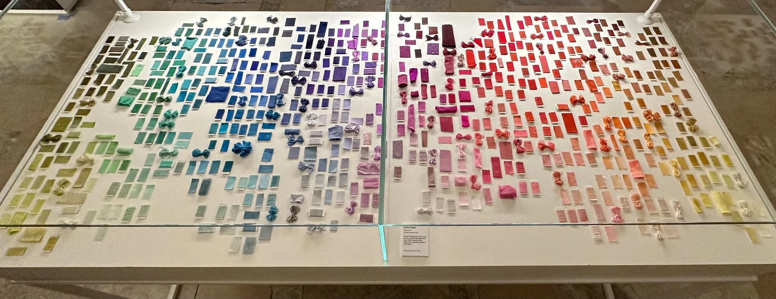 Rainbow-colored miniature objects arranged on white display table, organized by color from green to yellow, then blue, purple, pink, red, and orange, with a rail dividing the display into two sections.