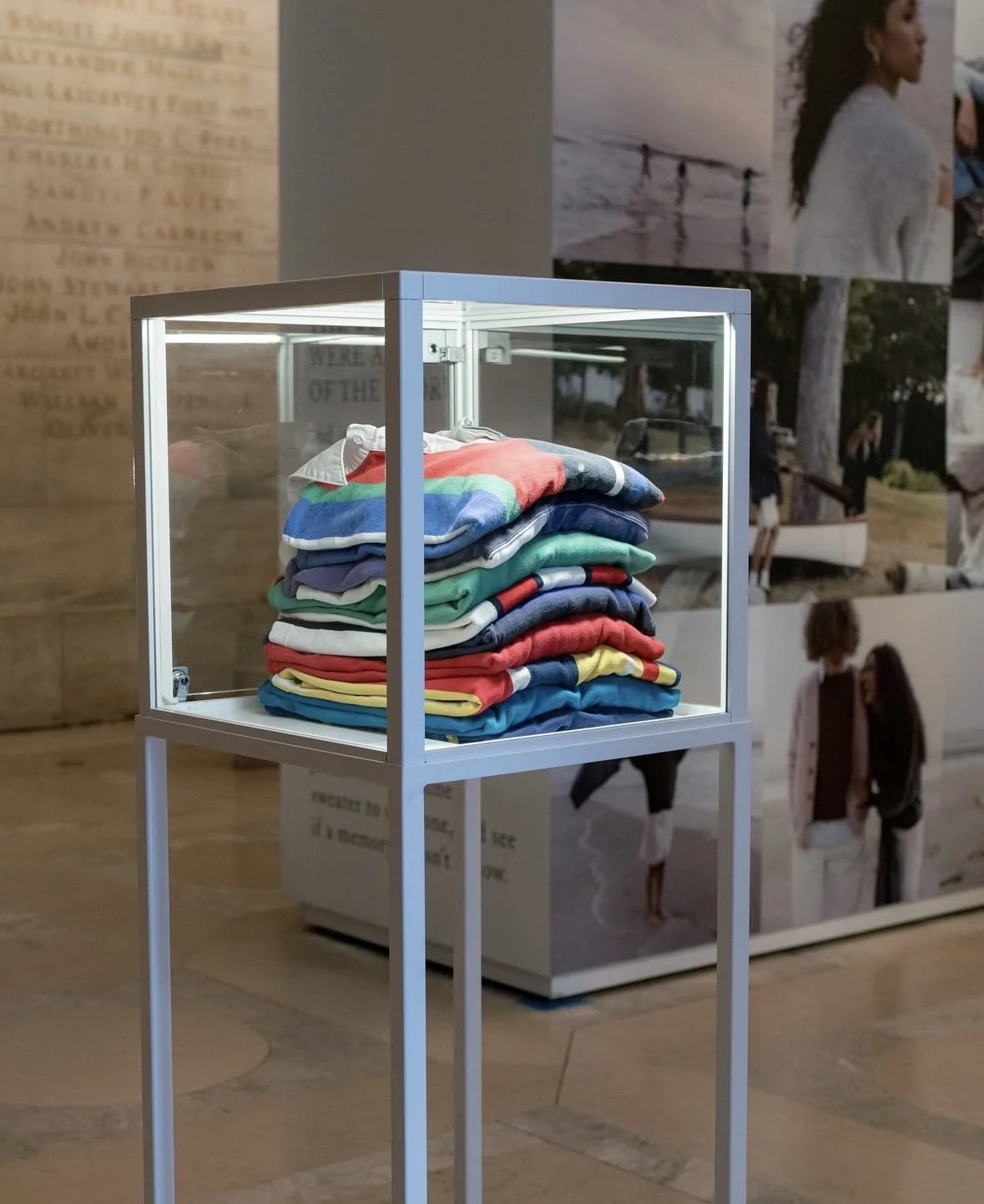 Display case with folded colorful t-shirts inside, located at an exhibition with photographic wall art in the background.